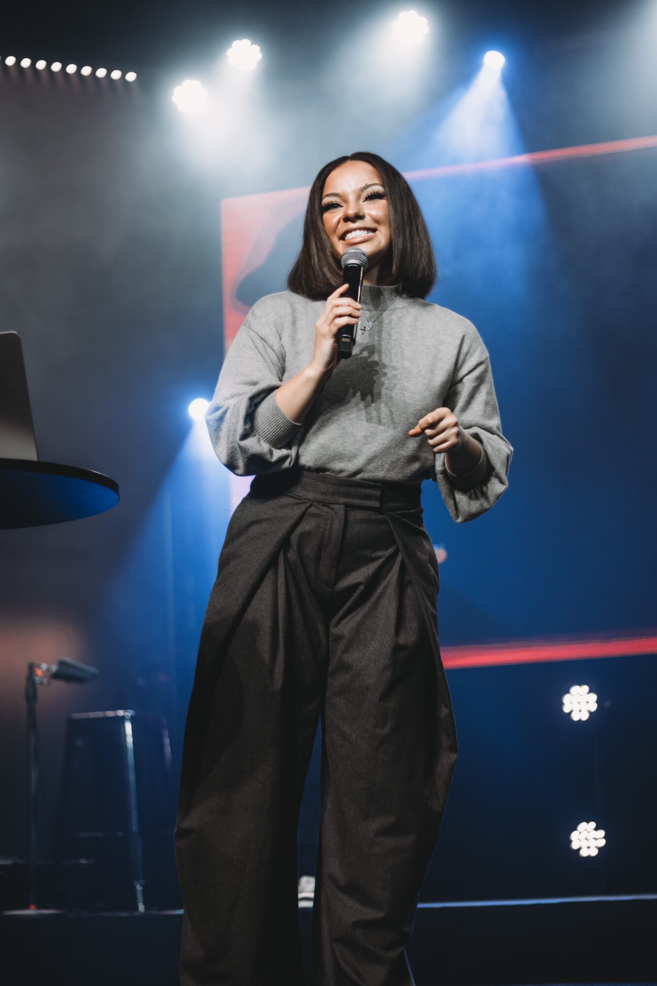 Woman with shoulder-length hair holding a microphone on stage, smiling, with stage lights in background.