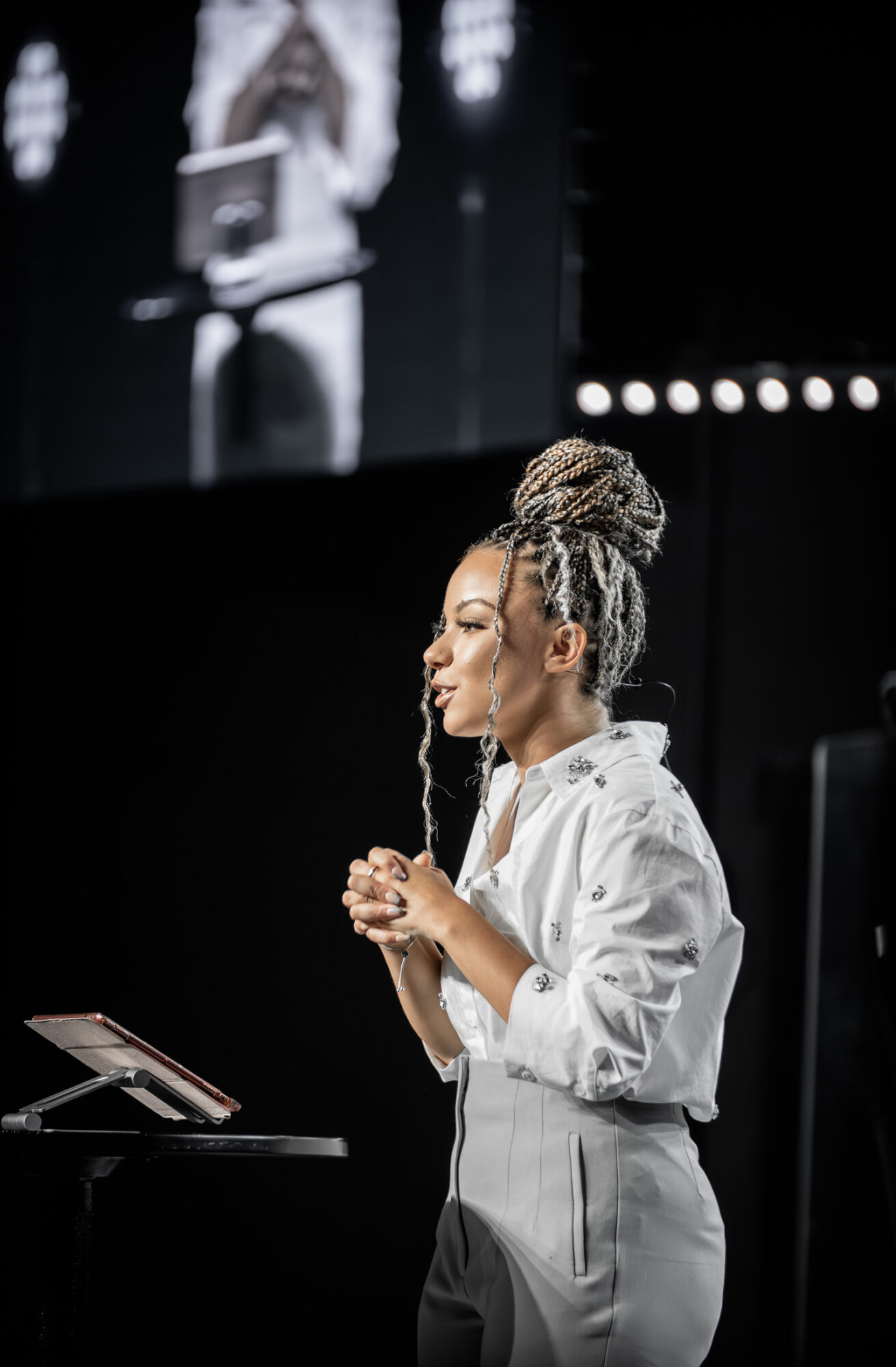 Woman with braided hair and white shirt speaking into a microphone on stage, with a large screen behind her.
