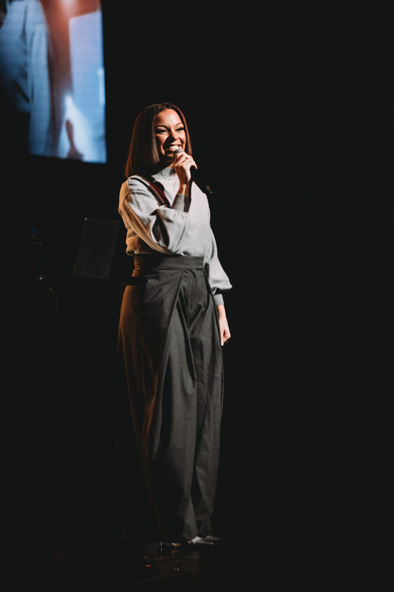 Woman standing on stage, smiling and holding a microphone, with a dark background and a screen behind her.