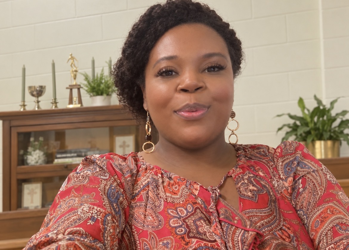Woman with short curly hair wearing a patterned red and gold top, smiling indoors with shelves and plants in background.