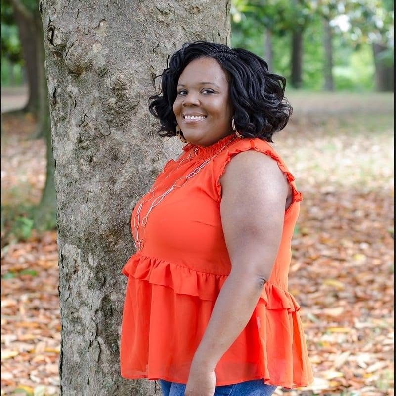 Woman with curly black hair smiling, wearing an orange sleeveless top, standing outdoors near a tree with fallen leaves.