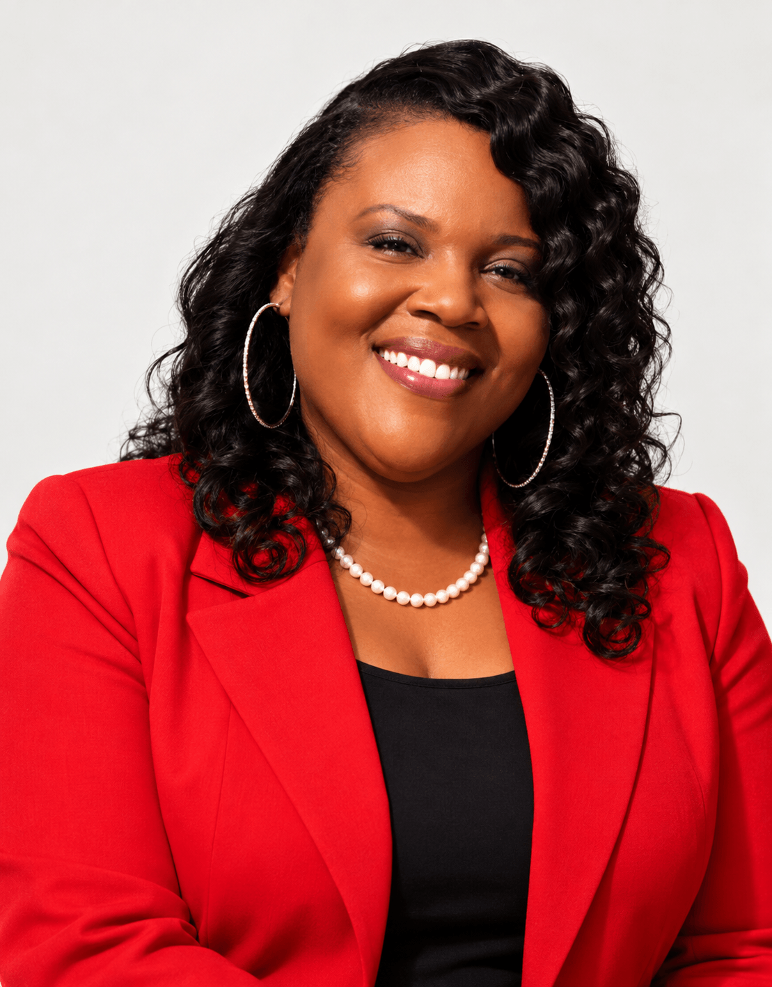 Smiling woman with curly hair, wearing a red blazer, black top, pearl necklace, and hoop earrings, against a white background.