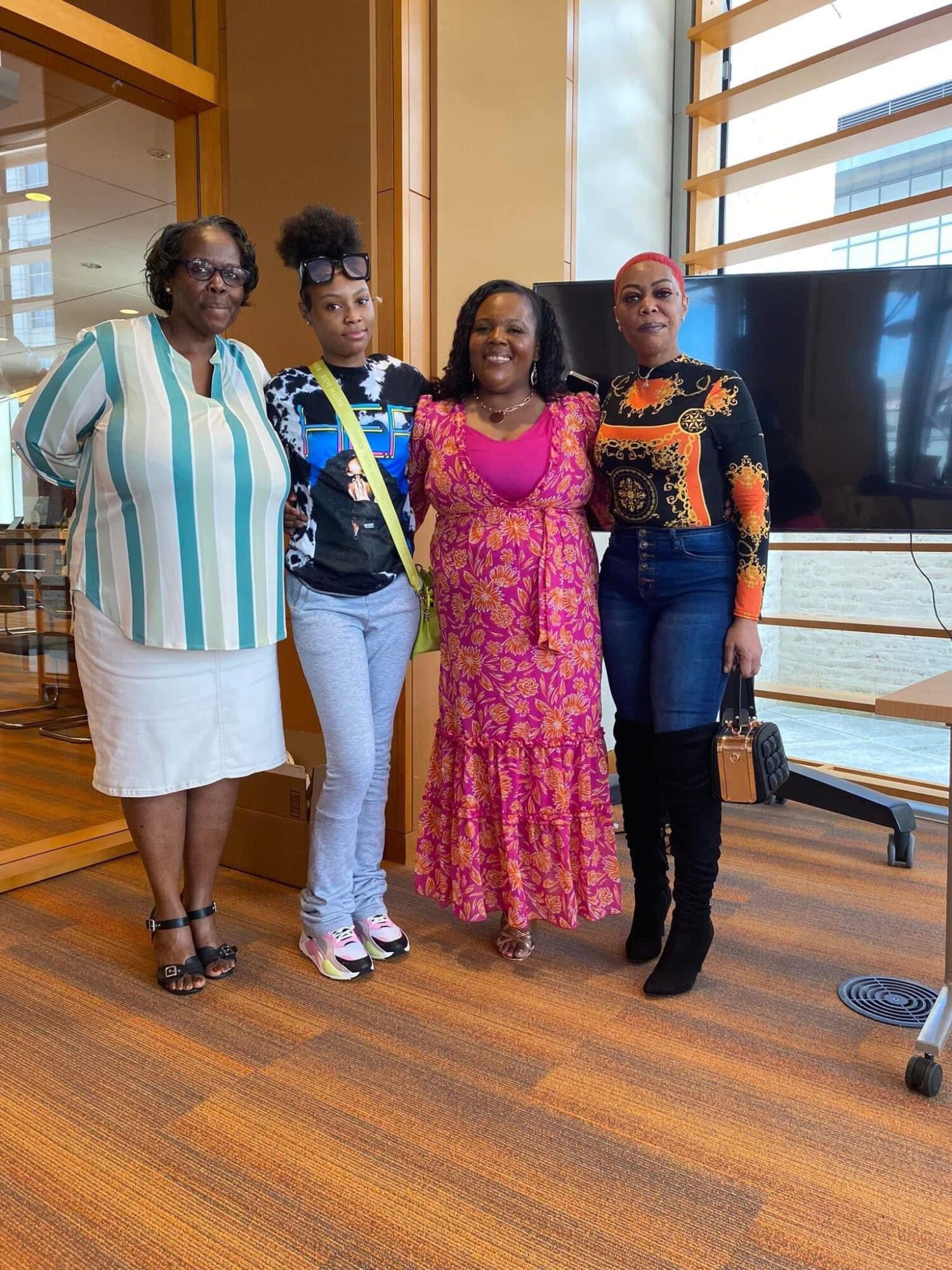 Four women standing indoors, smiling, with large windows and a television behind them.