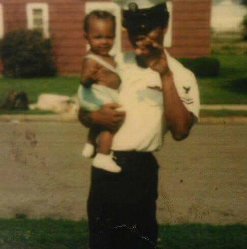 Person in uniform holding a smiling child outdoors, background with houses and greenery, sunny day.