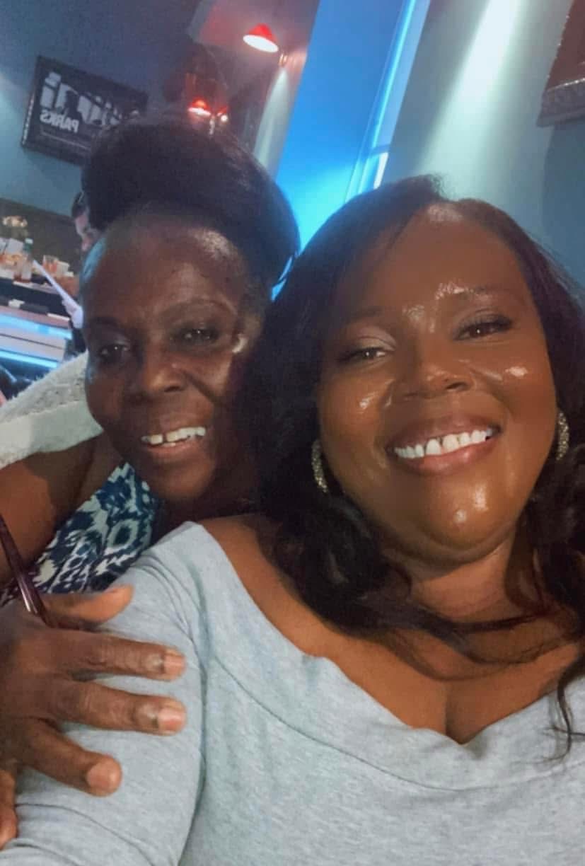 Two women smiling and posing for a selfie indoors, with colorful lighting in the background.