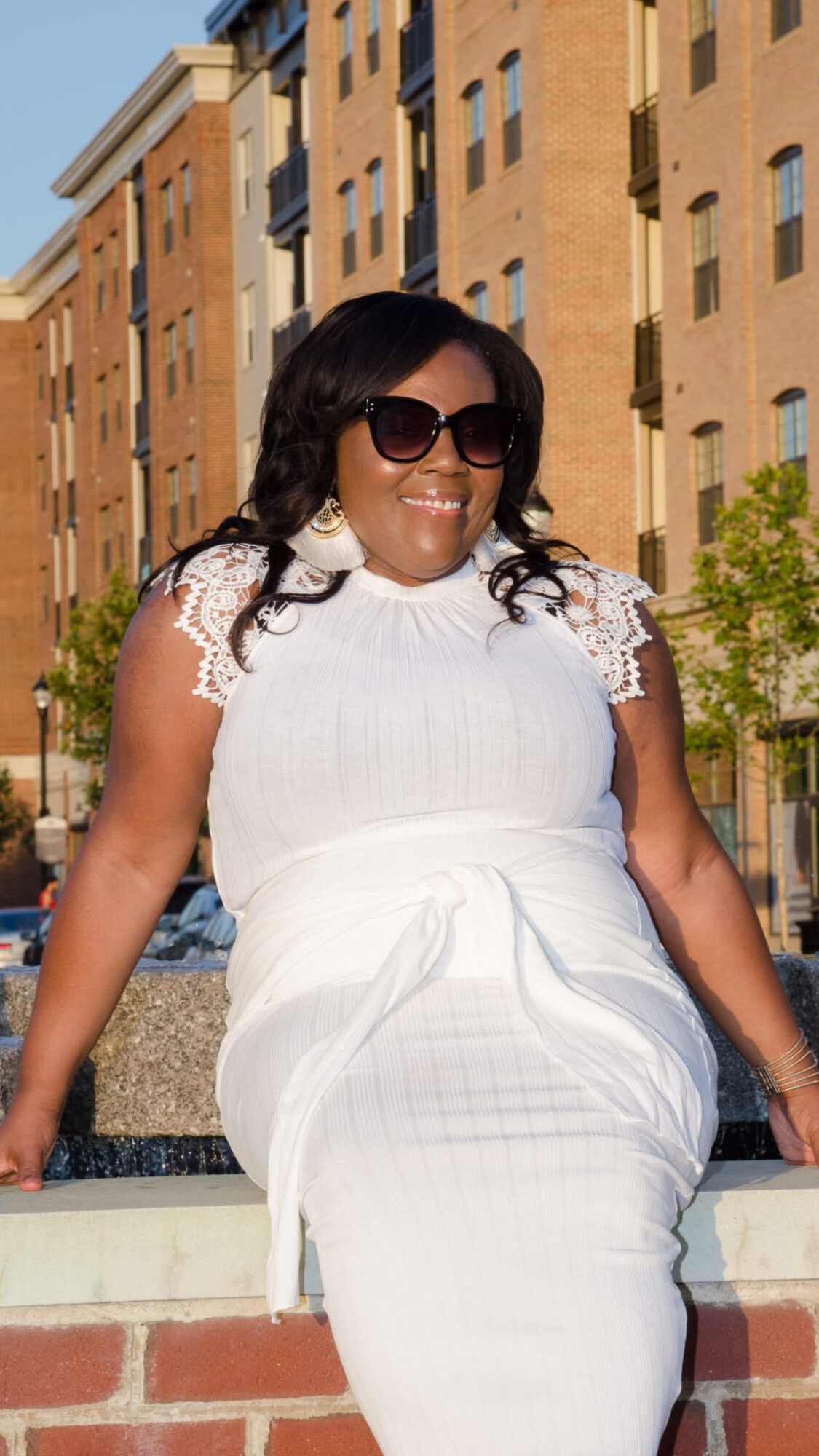 Woman sitting outdoors on a ledge, wearing sunglasses and a white dress, smiling, with buildings in the background.