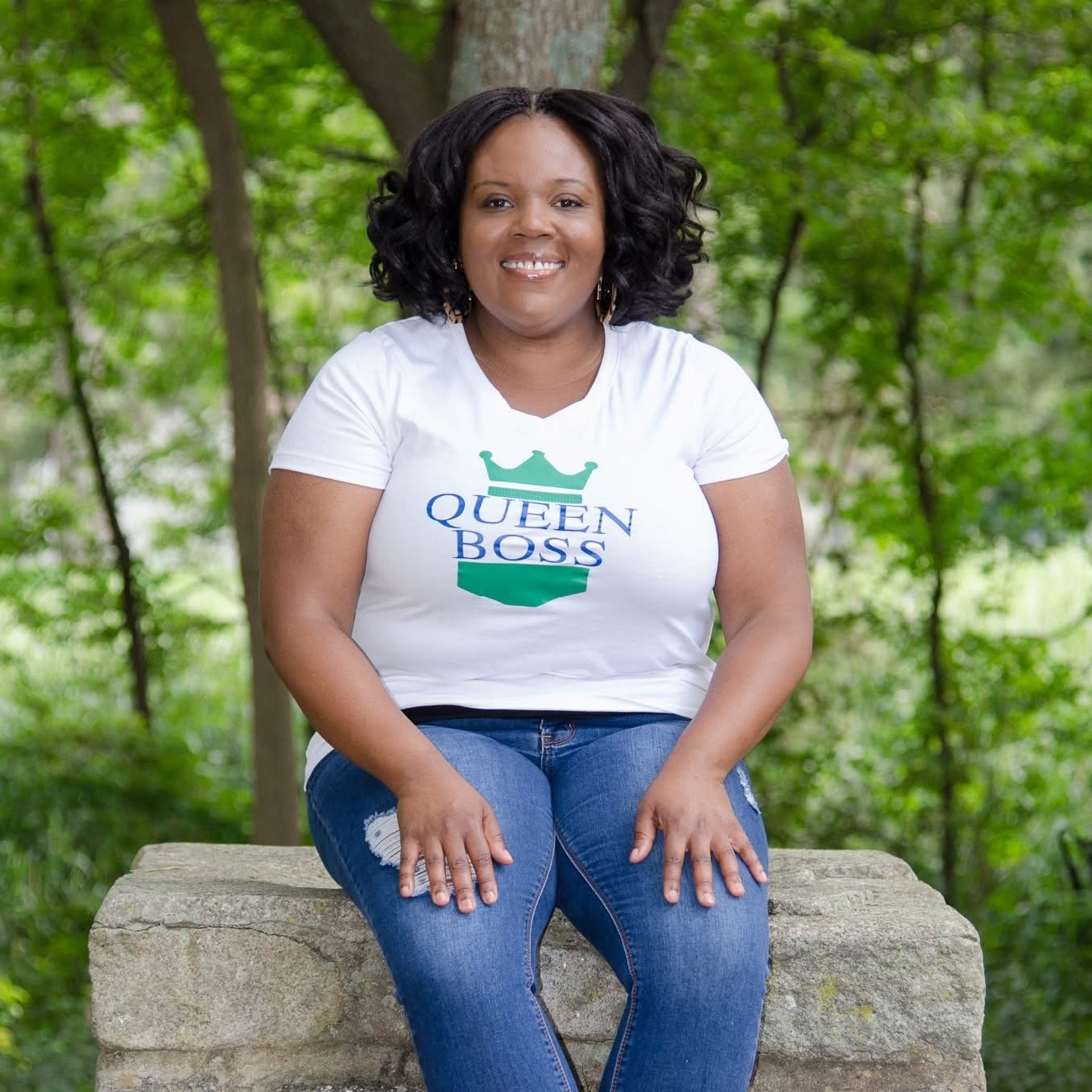 Woman sitting on a stone bench outdoors, smiling, wearing a white T-shirt and blue jeans, surrounded by green trees.