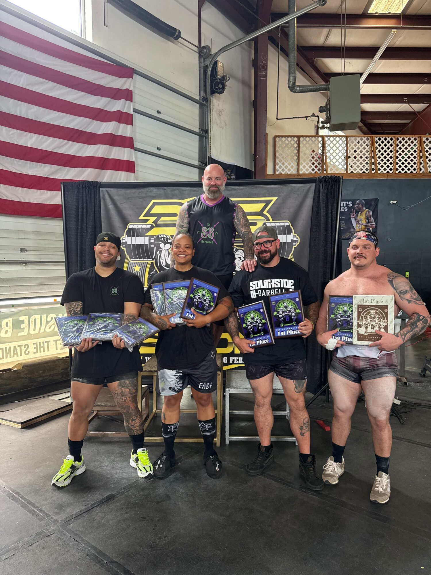 Group of five people holding gaming consoles and trophies, standing in a warehouse or garage setting with an American flag and a black banner in the background.