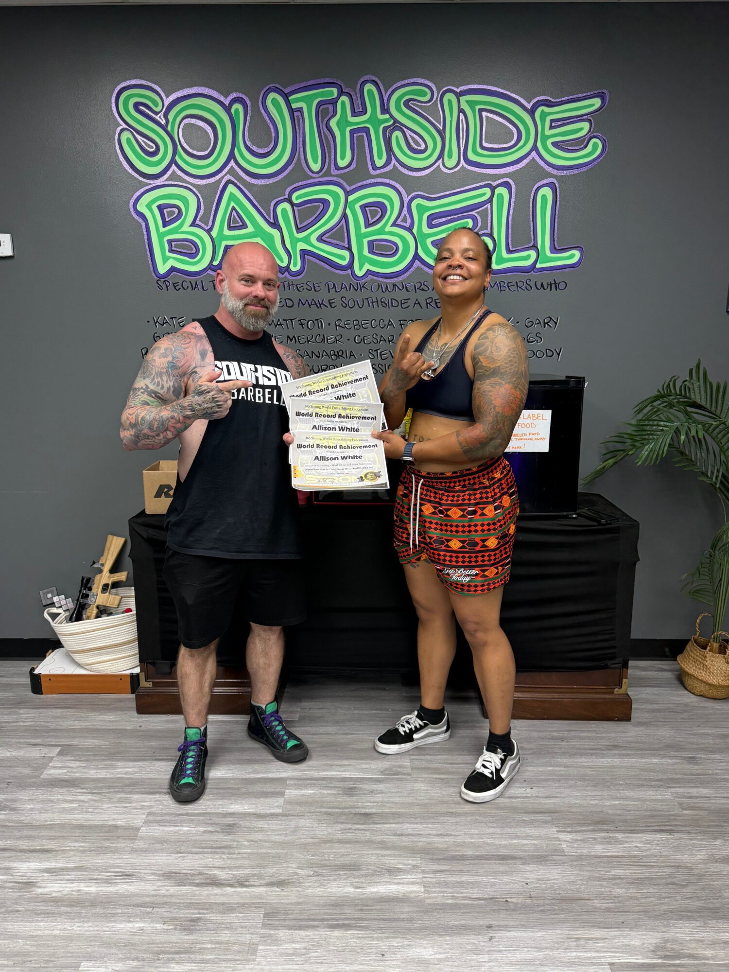 Two people standing in front of a gray wall with colorful text, one holding a certificate, the other with tattoos, both smiling.