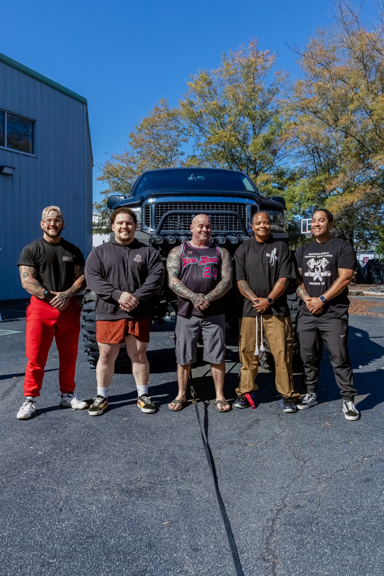 Five people stand outdoors in front of a large truck, with trees and a building in the background, on a sunny day.