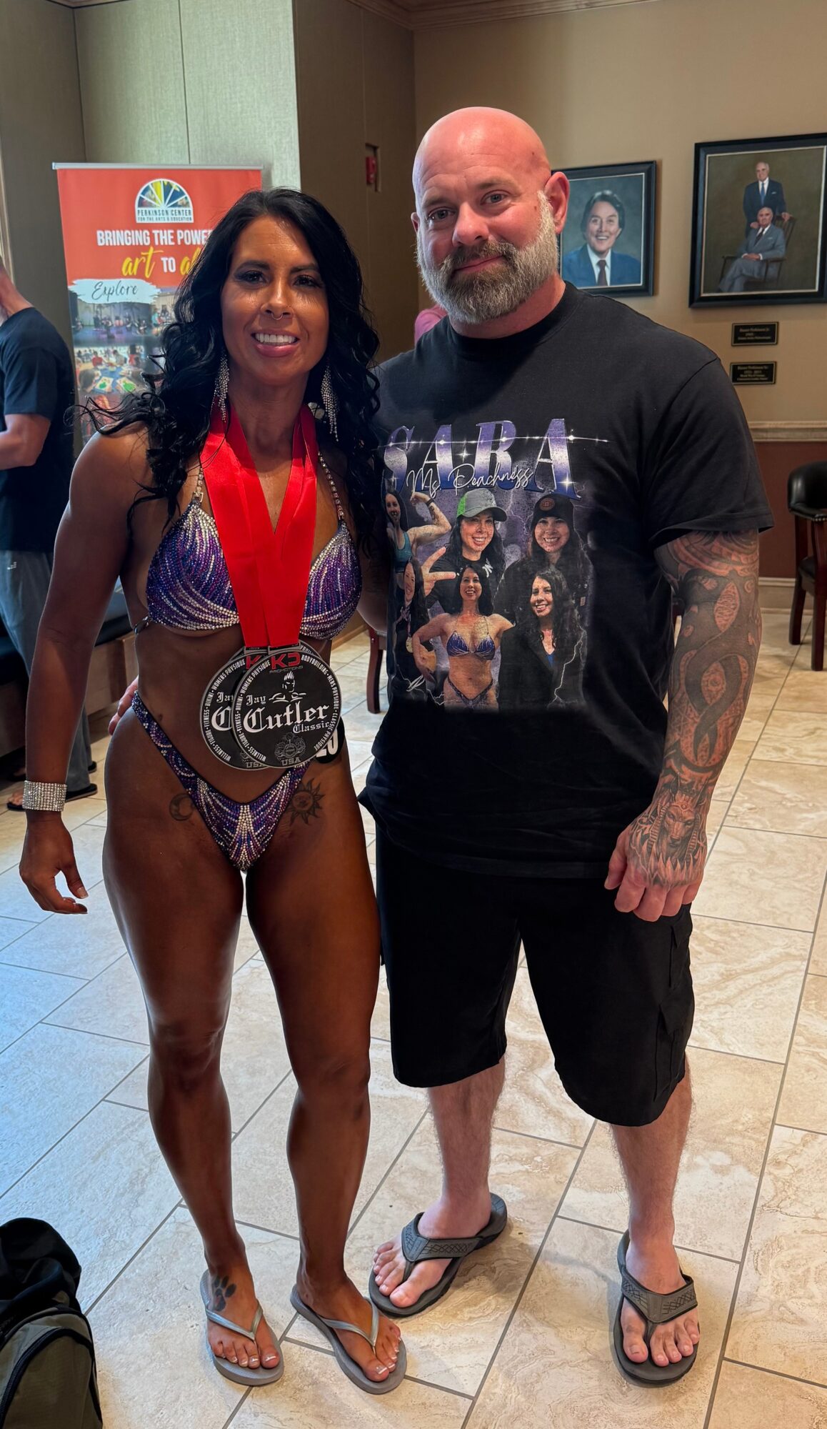 A woman in a bikini with a medal and a man in casual clothing stand together indoors, smiling.