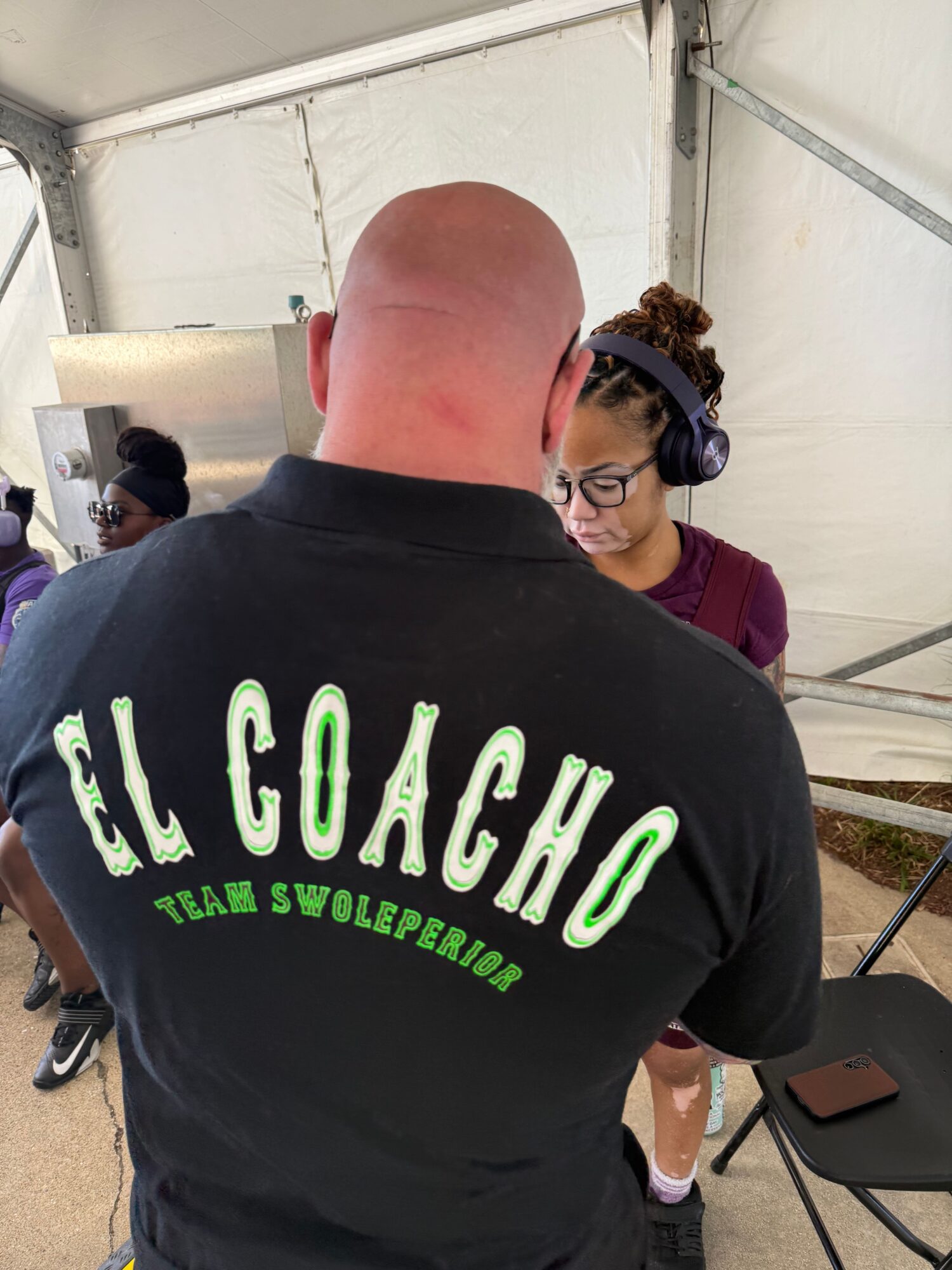 Bald man with a black shirt talking to a woman with glasses and headphones inside a tent.