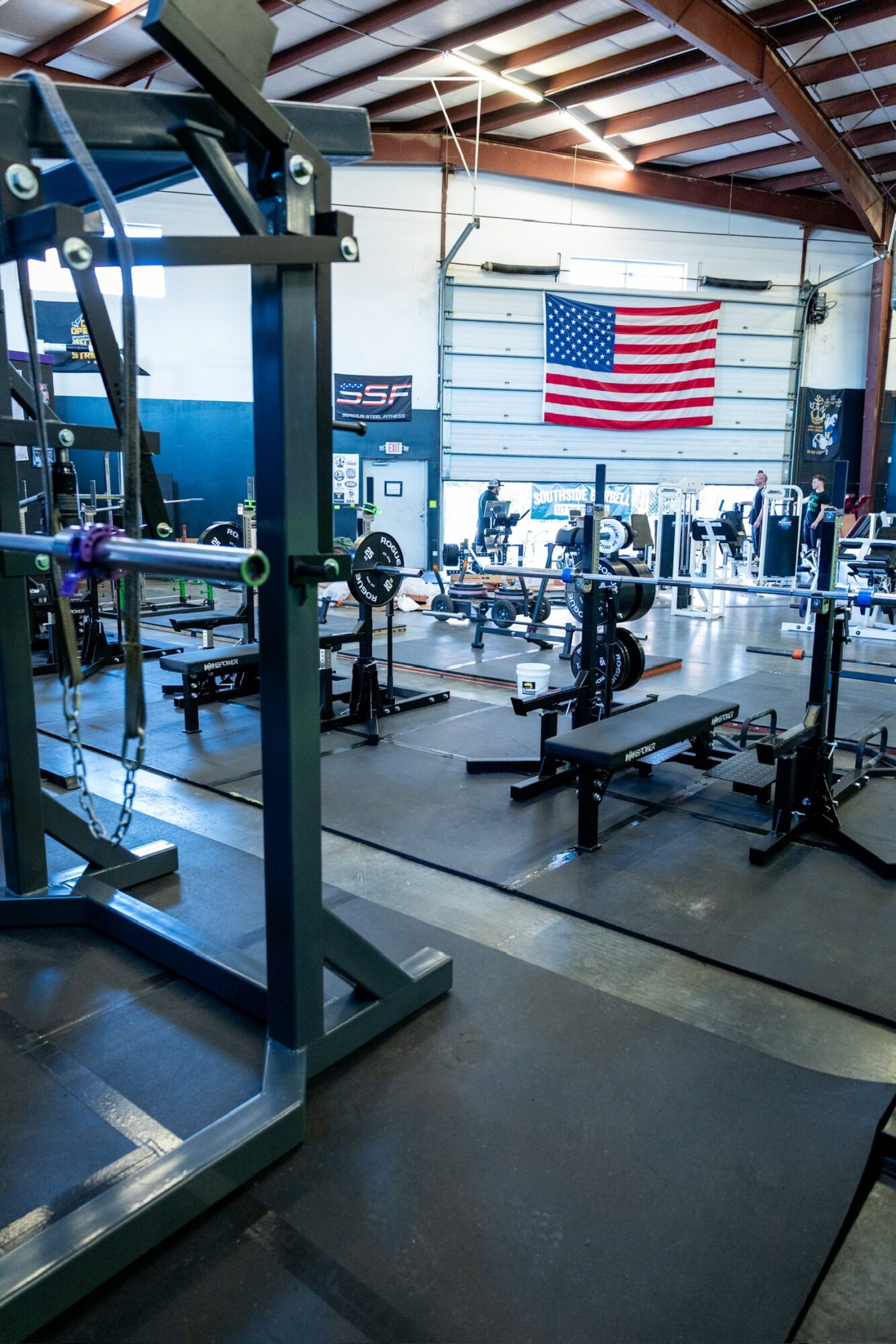Gym with weightlifting equipment and an American flag on the wall, inside a spacious warehouse-style building.
