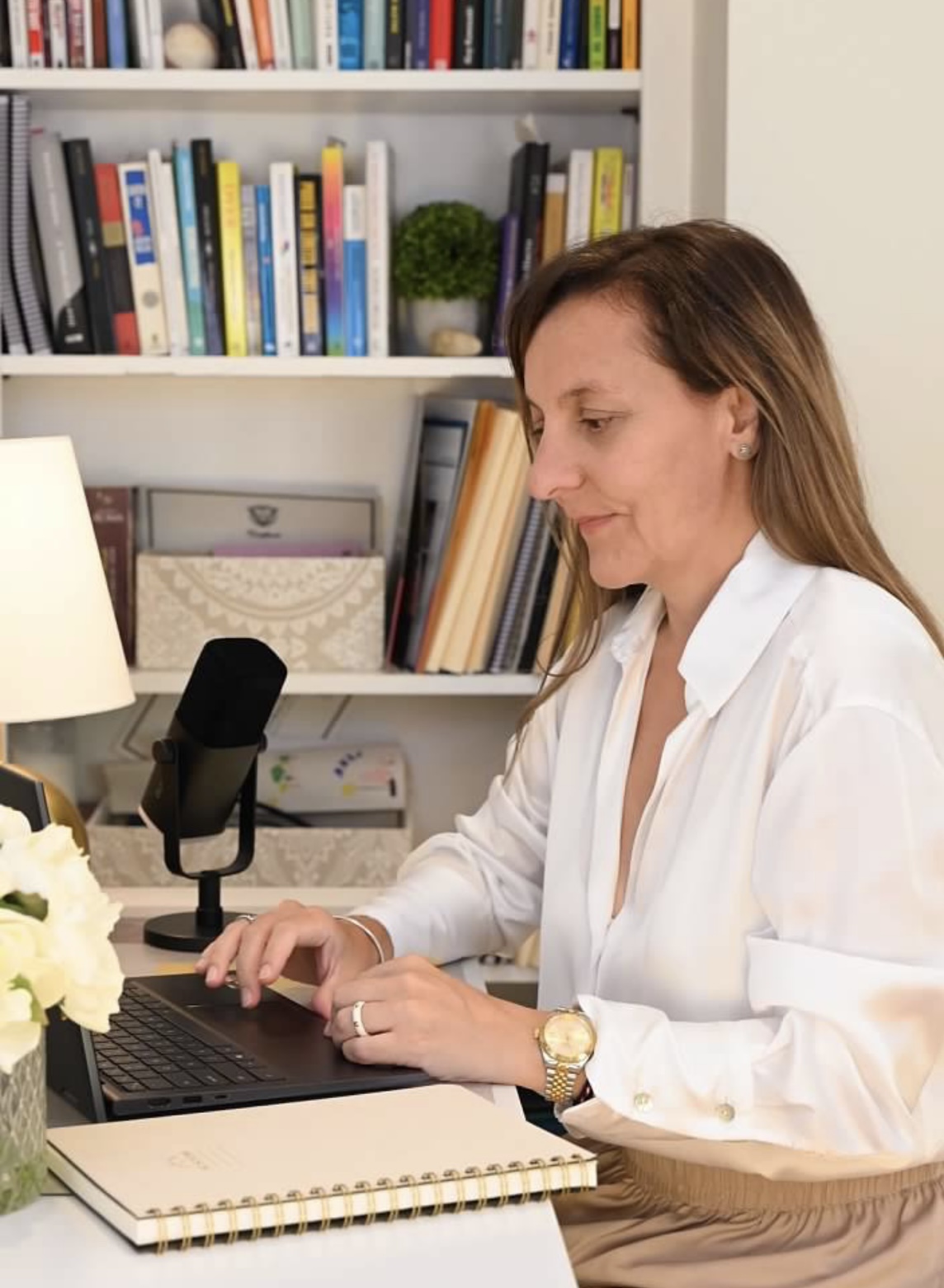 Woman sitting at desk with laptop, bookshelves in background, wearing white shirt, smiling, with a microphone nearby.