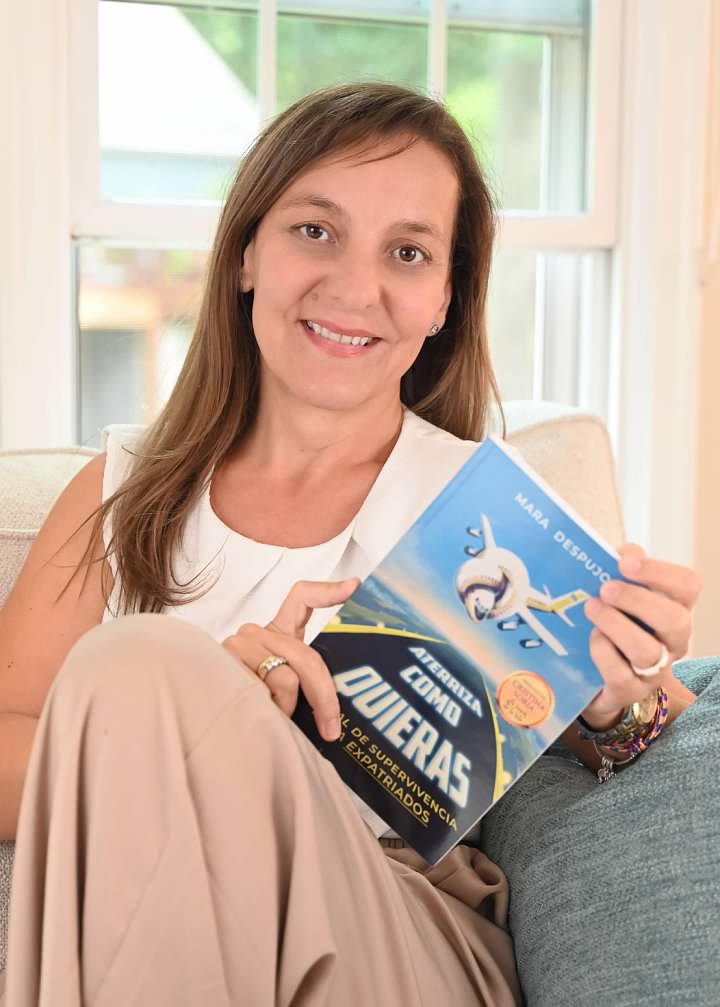 Woman sitting on a couch holding a book titled 'Queridas' with a white airplane illustration on the cover, smiling at the camera.