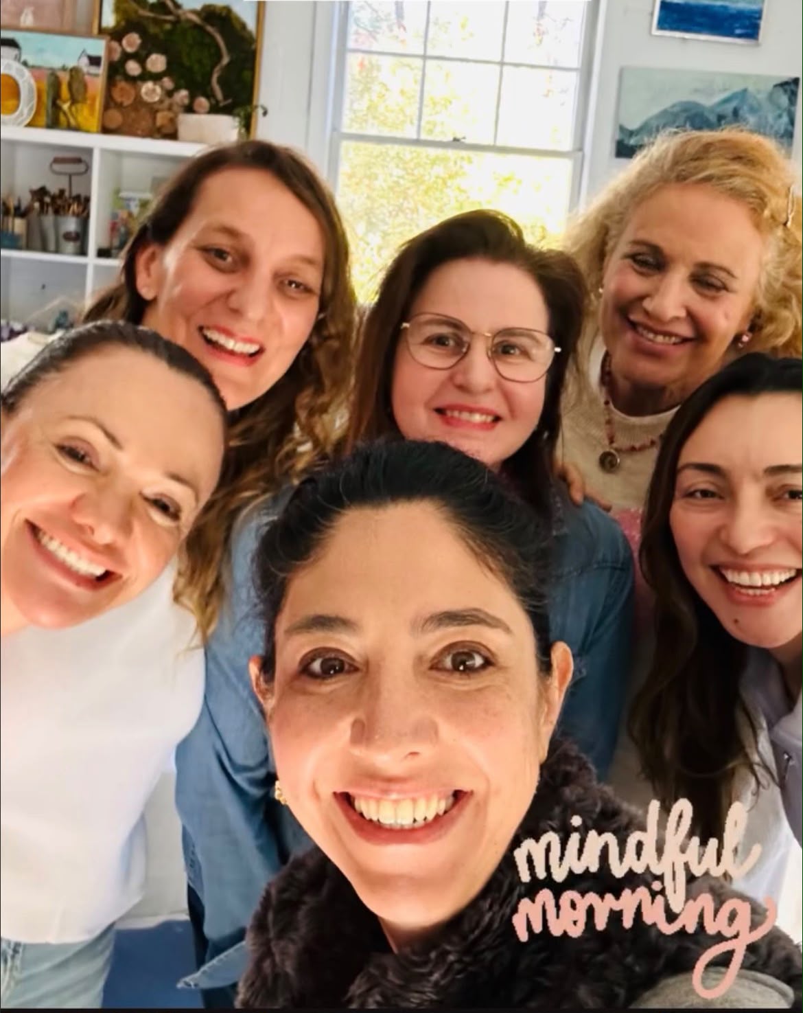 Six women smiling closely together indoors with a window and artwork in the background.