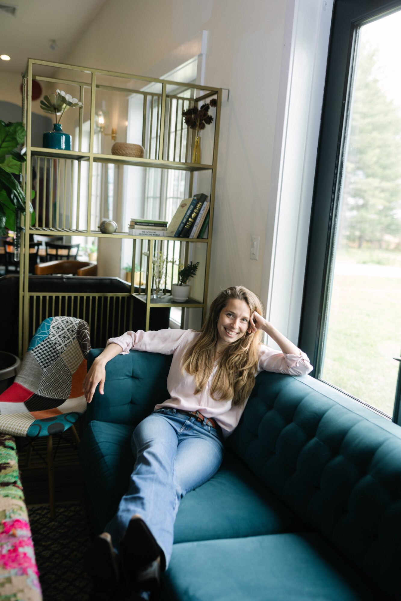 Young woman with long hair sitting on a teal sofa near a large window, smiling and resting her head on her hand.