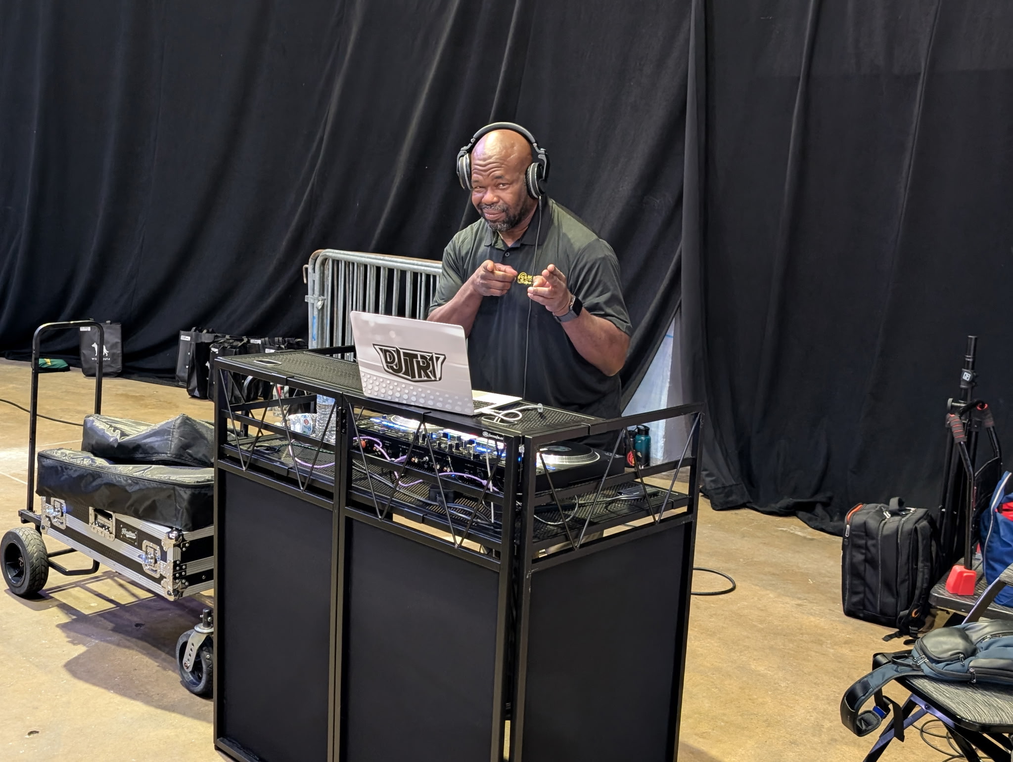 DJ wearing headphones stands behind equipment with a laptop, in front of black curtains, holding a snack or object.