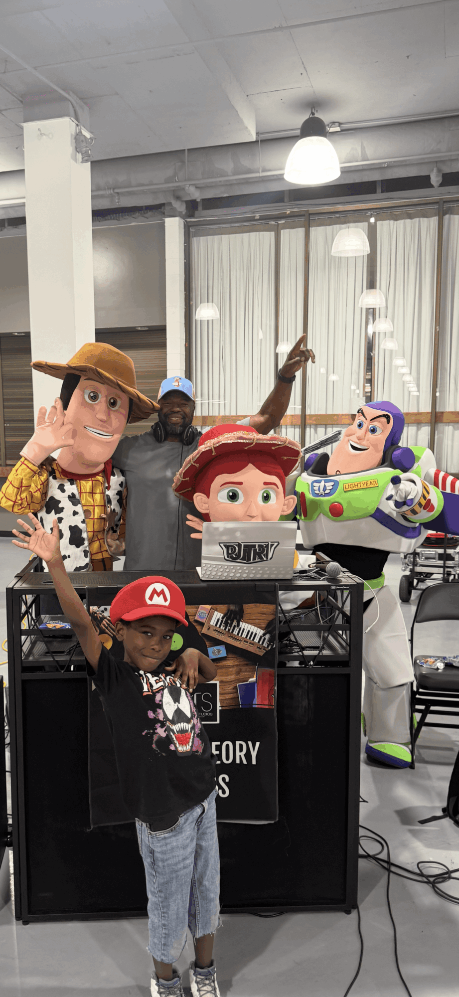 Children with Toy Story characters in a room with large windows and hanging lights, some children wearing hats and a Mario shirt.