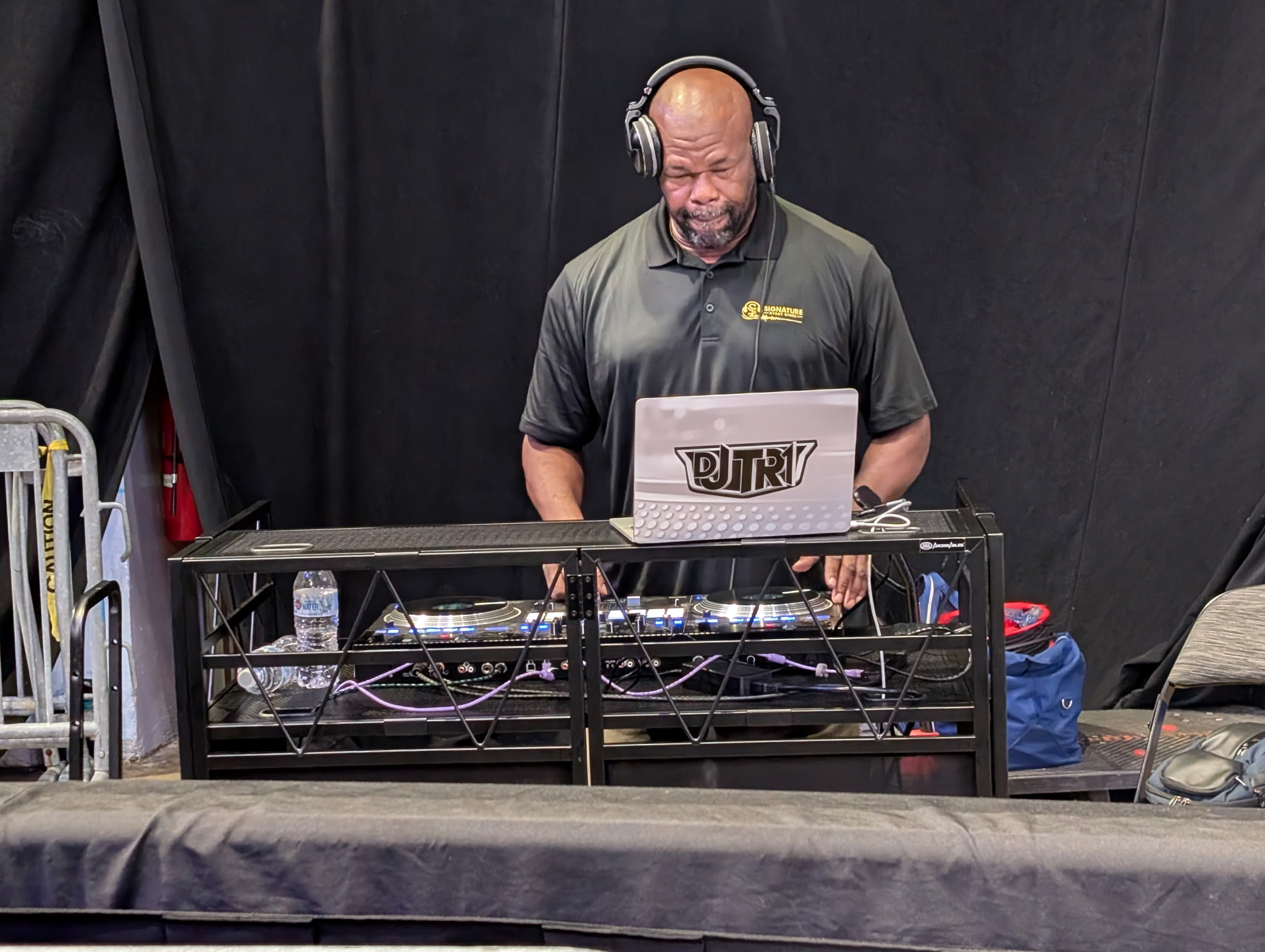 DJ wearing headphones stands behind a DJ setup with a laptop, in front of a black backdrop.