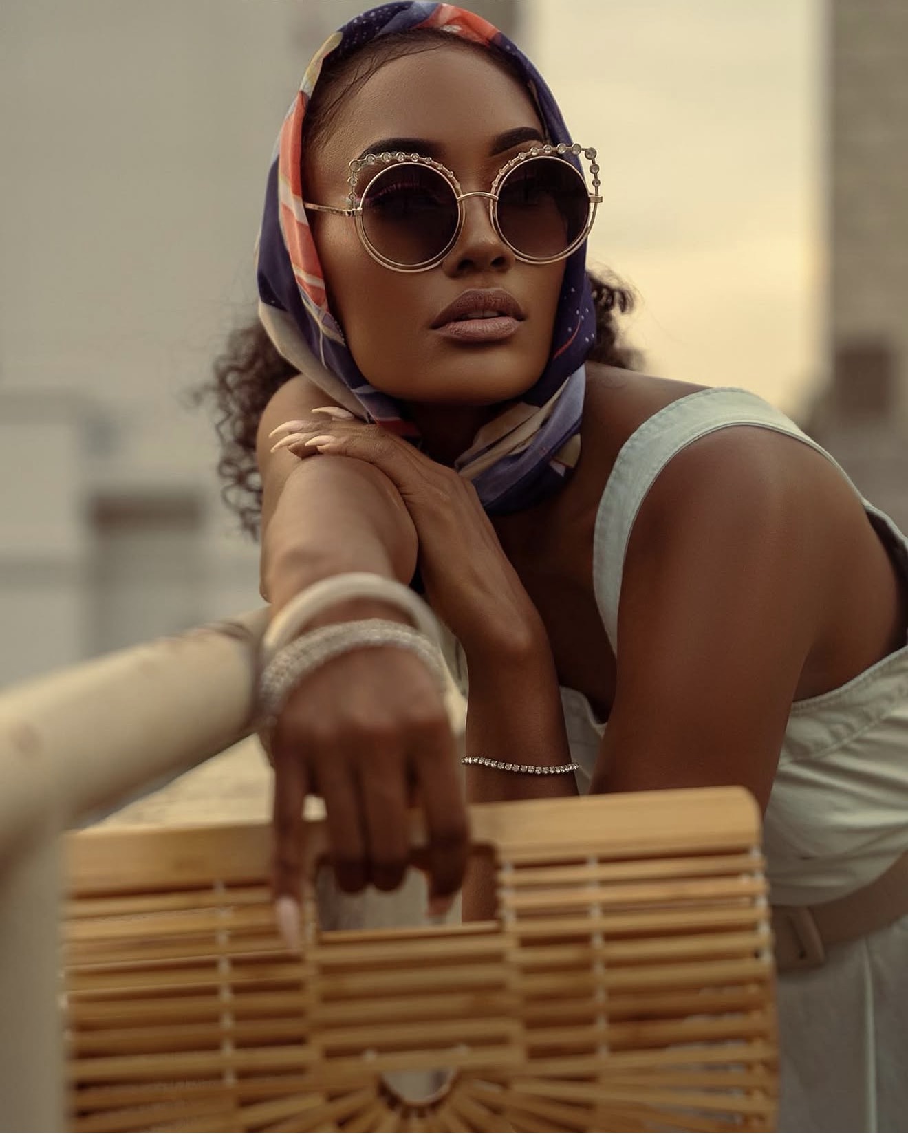 Woman wearing large sunglasses and a headscarf, resting her chin on her hand, holding a woven bag, outdoors.