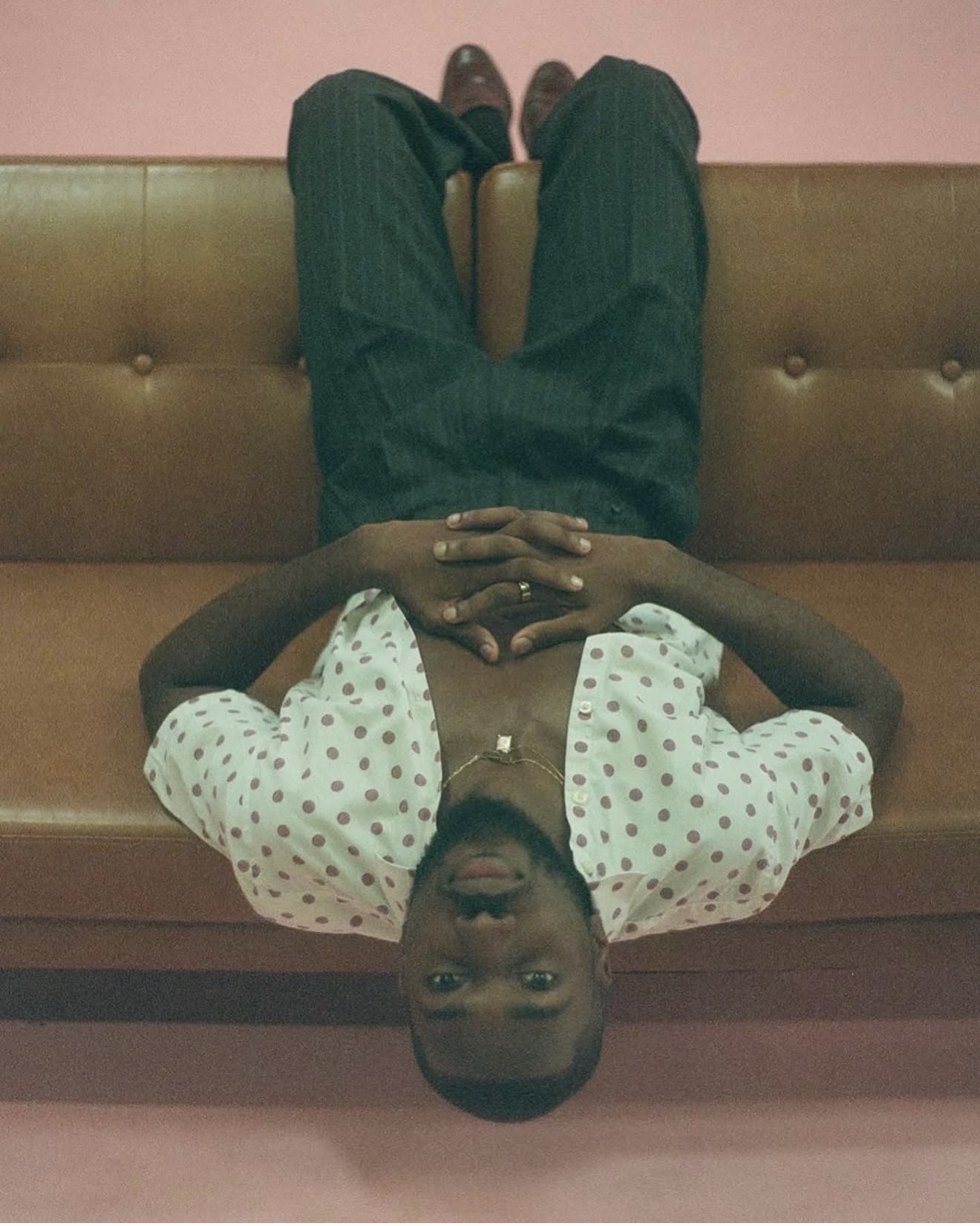 Person lying on a couch with legs up, wearing a polka dot shirt and dark pants, looking at the camera.