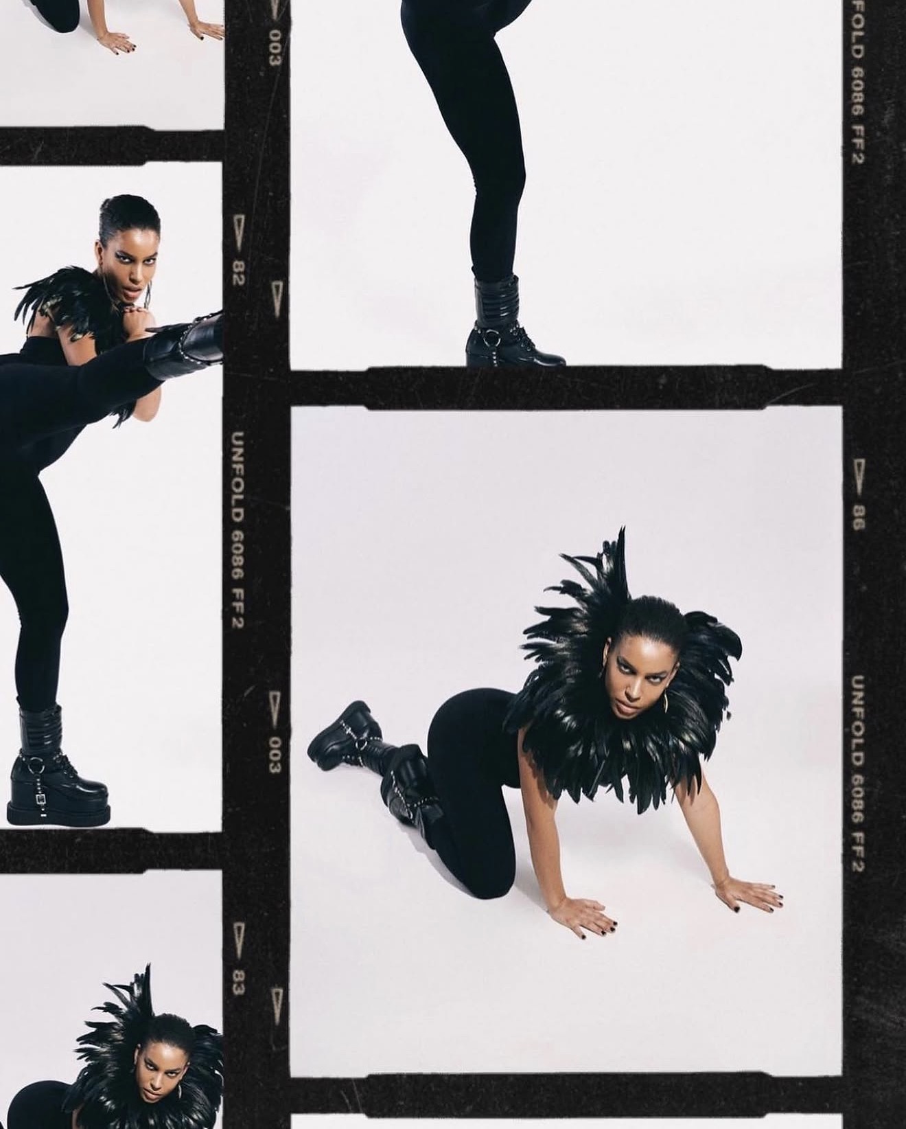 Person with long hair and feathered costume crouches on the floor, with another person's legs visible above, against a white background.