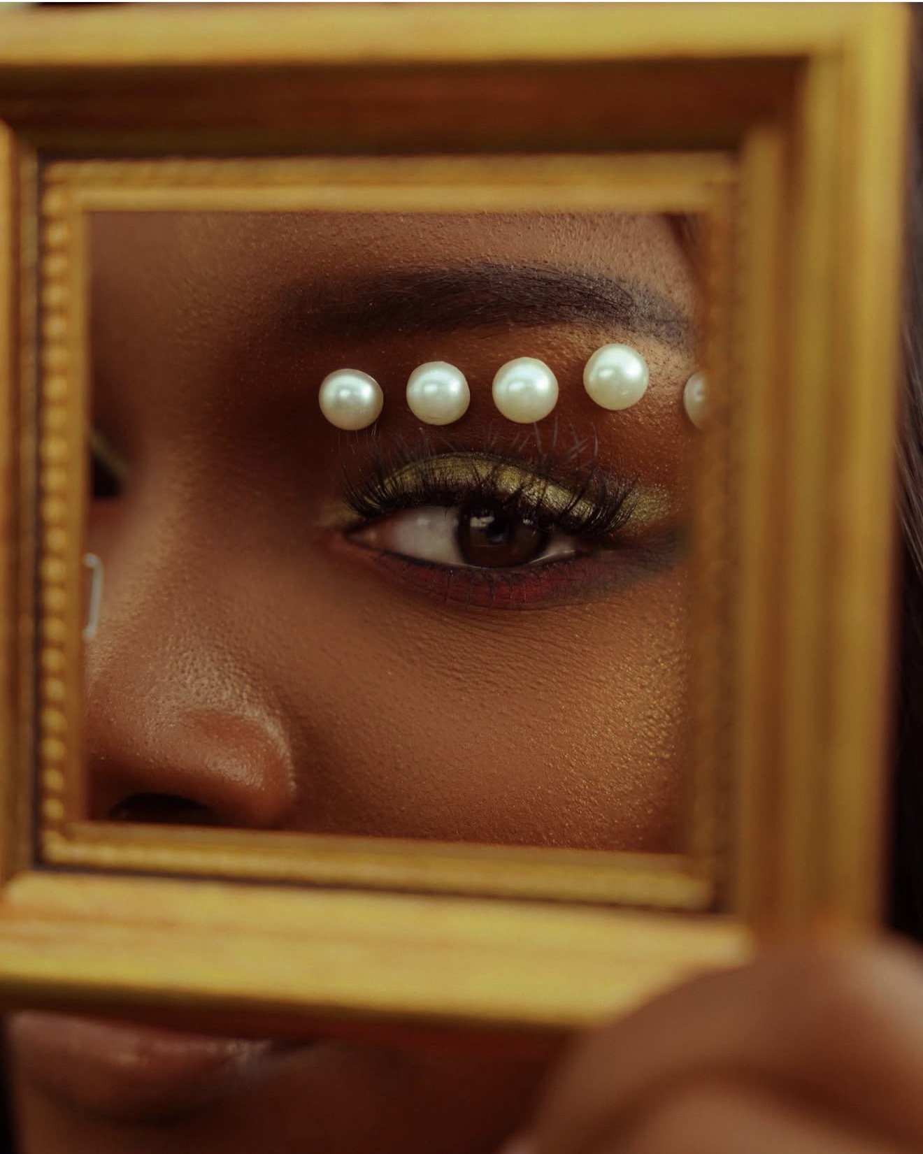 Close-up of a person's eye with pearl embellishments above the eyebrow, reflected in a small wooden mirror.