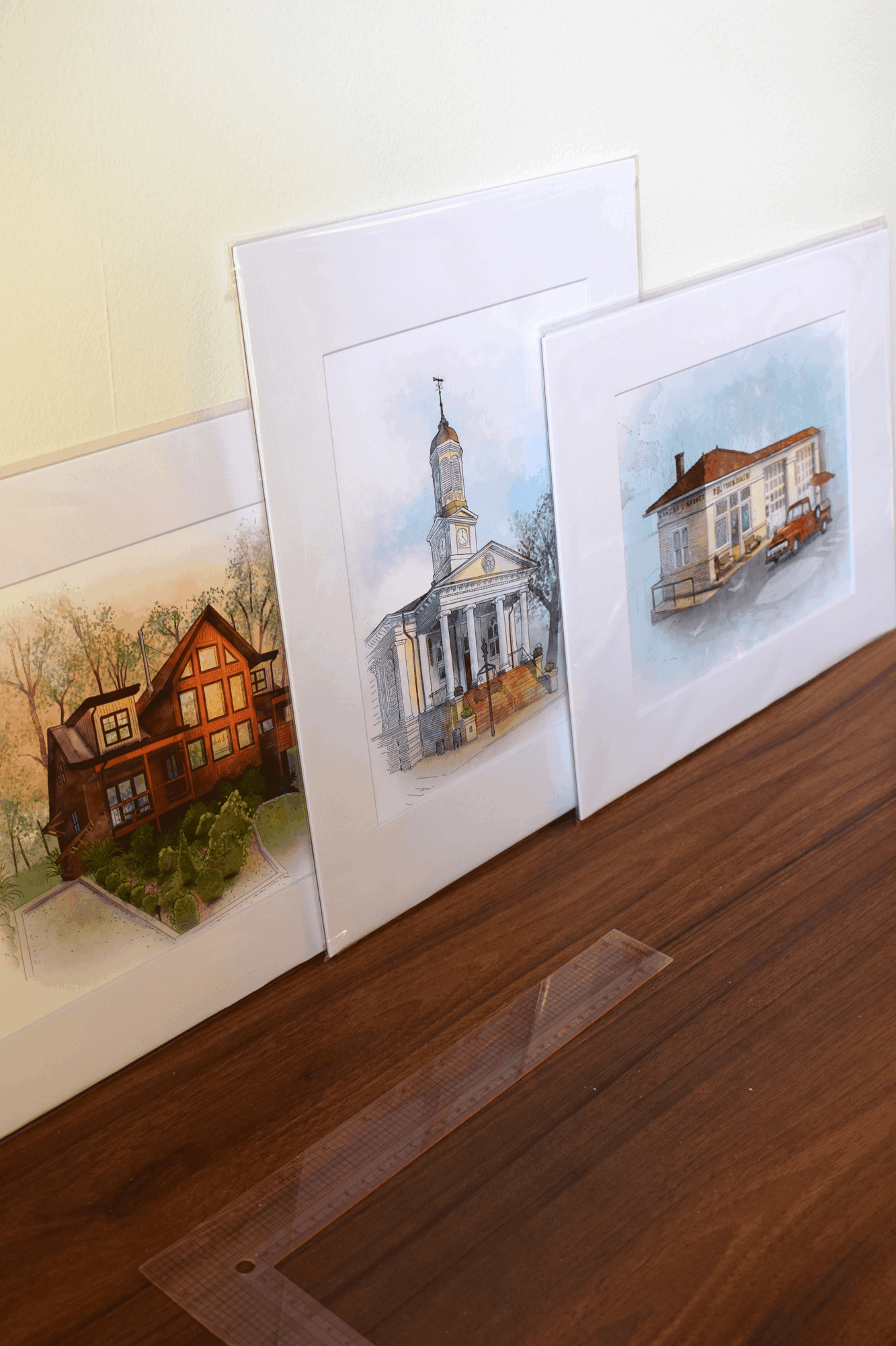 Four watercolor paintings of houses and buildings leaning against a wall on a wooden surface.