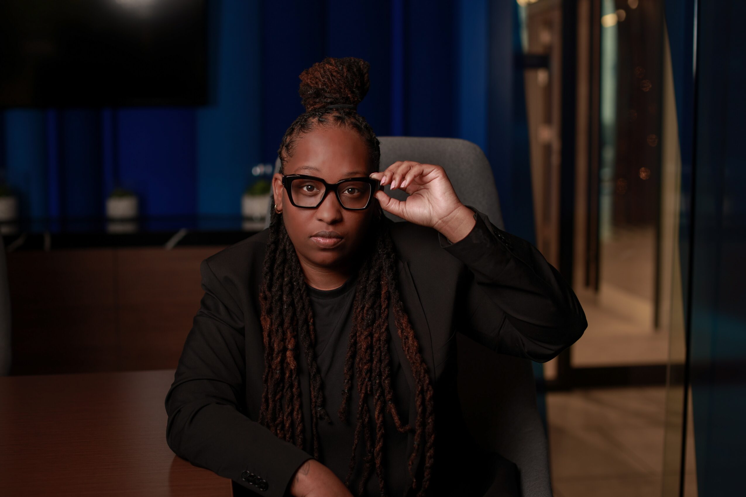 Woman with glasses and braided hair sitting in a chair, adjusting her glasses, in a modern indoor setting.