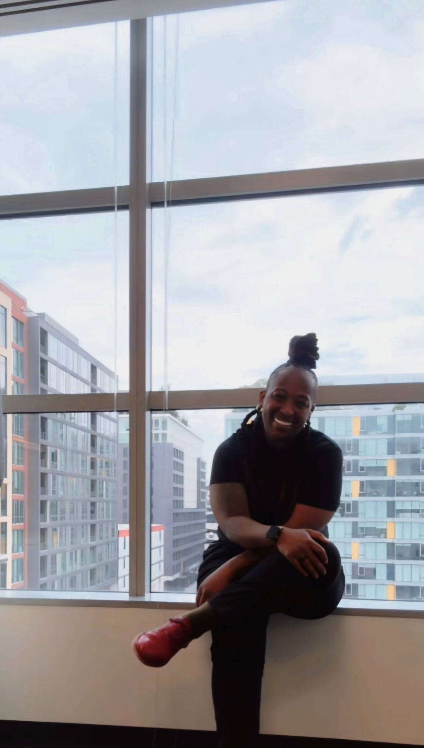 Person sitting on window ledge smiling, city buildings visible outside large window, daytime.