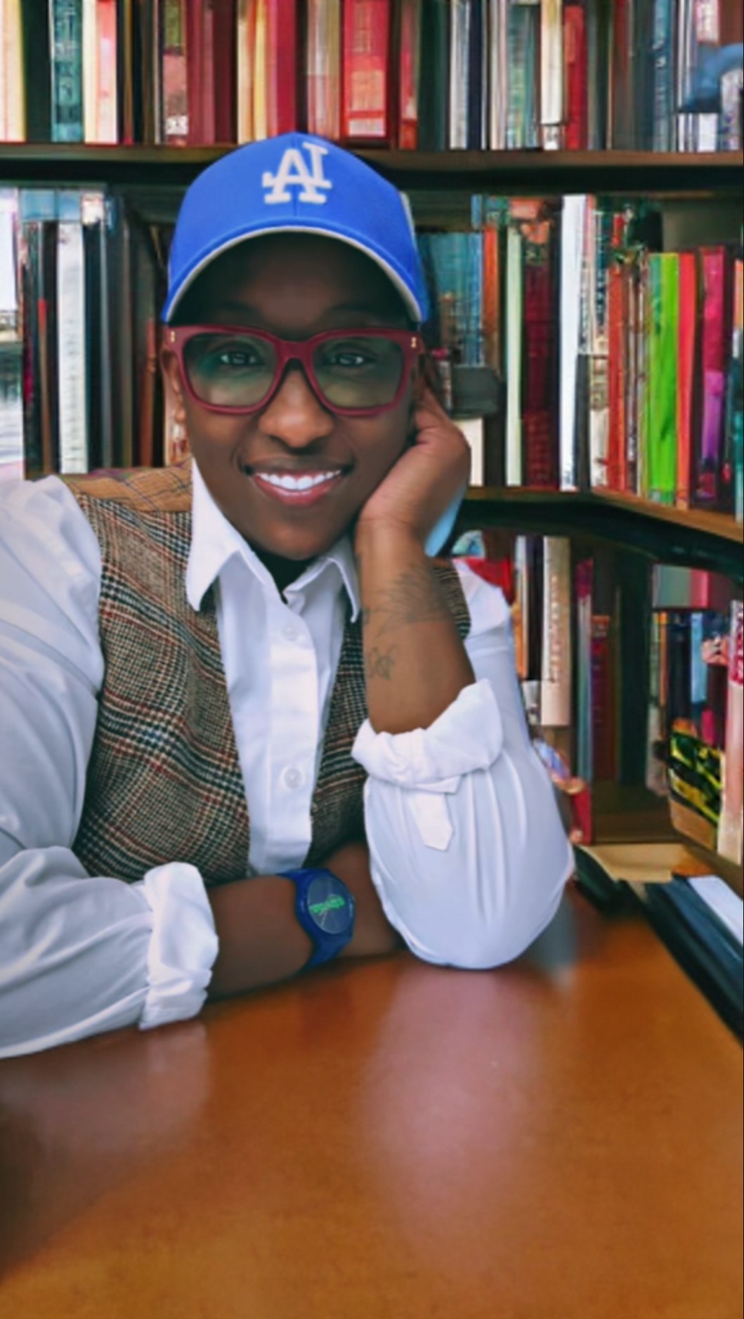 Person wearing a blue cap and glasses, smiling, sitting at a table in front of bookshelves.