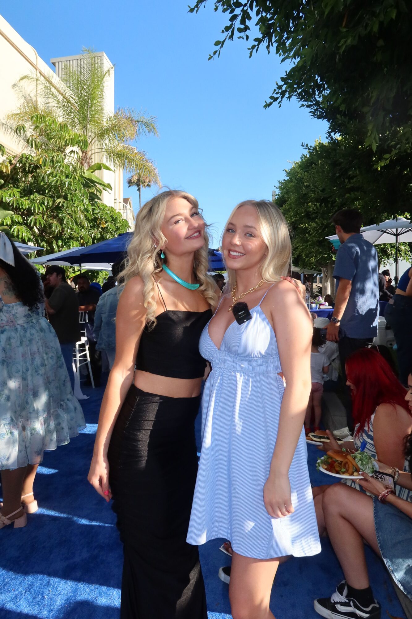 Two young women standing outdoors on a sunny day, one in a white dress and the other in black, with trees and a blue sky in the background.