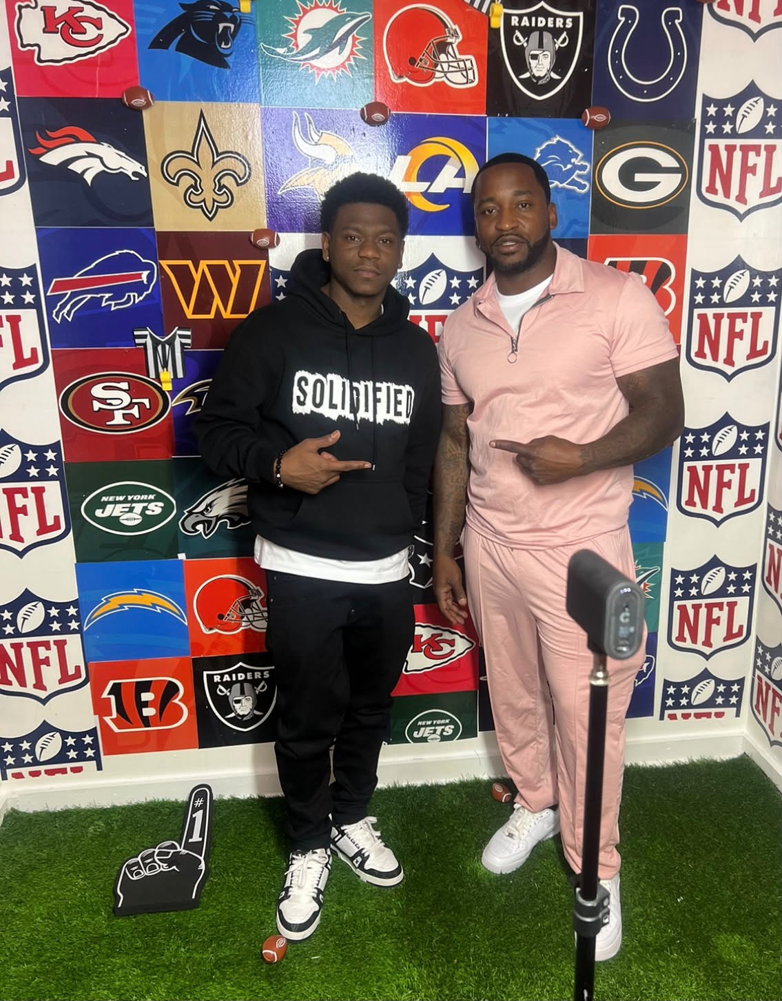 Two men standing in front of a wall decorated with NFL team logos, one pointing at the other, on a green carpeted floor.