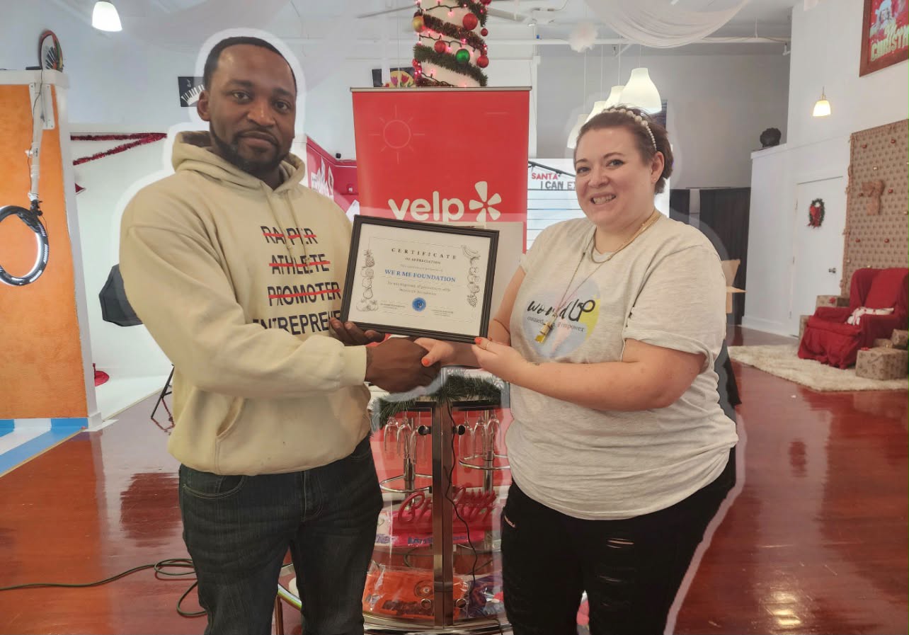 Two people holding a framed certificate together indoors, with a red Yelp banner behind them, smiling at the camera.