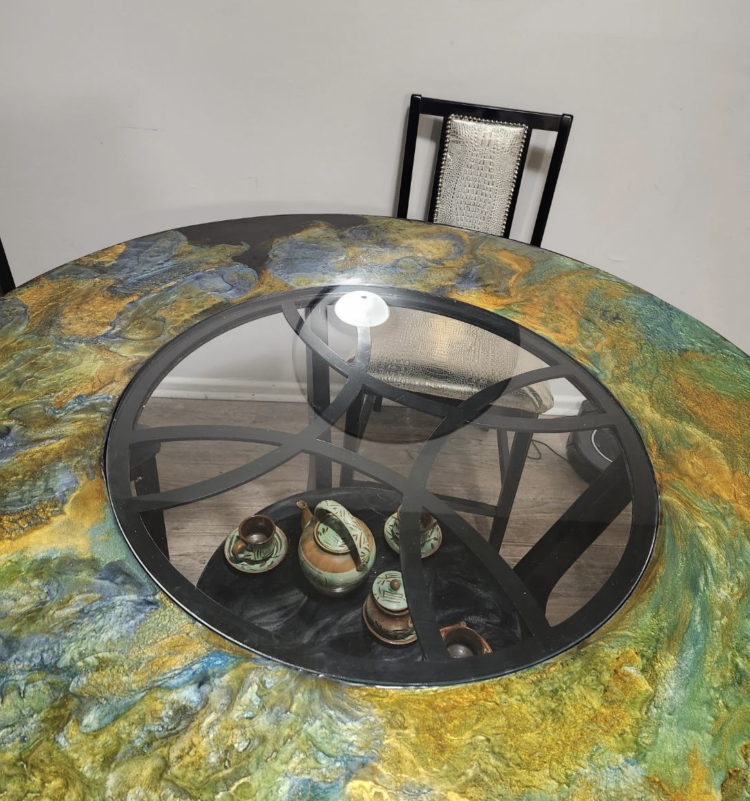 Round table with a colorful, textured surface and a glass center, surrounded by chairs, one with a black frame and woven backrest.