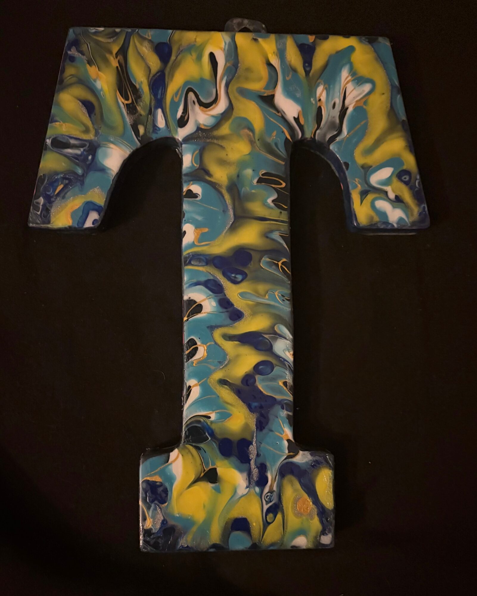 Letter T with a colorful marbled pattern in yellow, blue, white, and black on a dark background.
