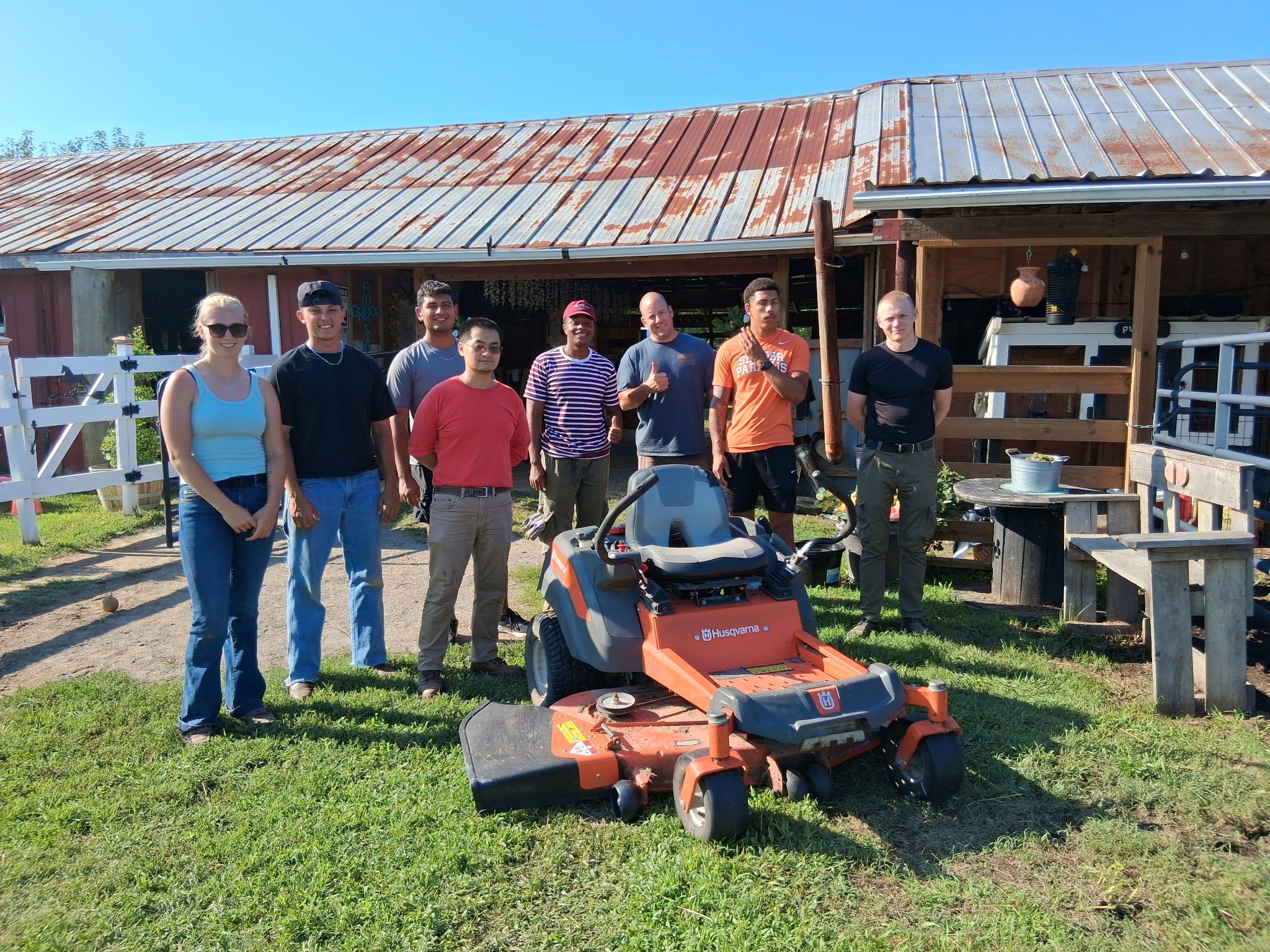 A group of people stands behind an orange riding mower in front of a barn.