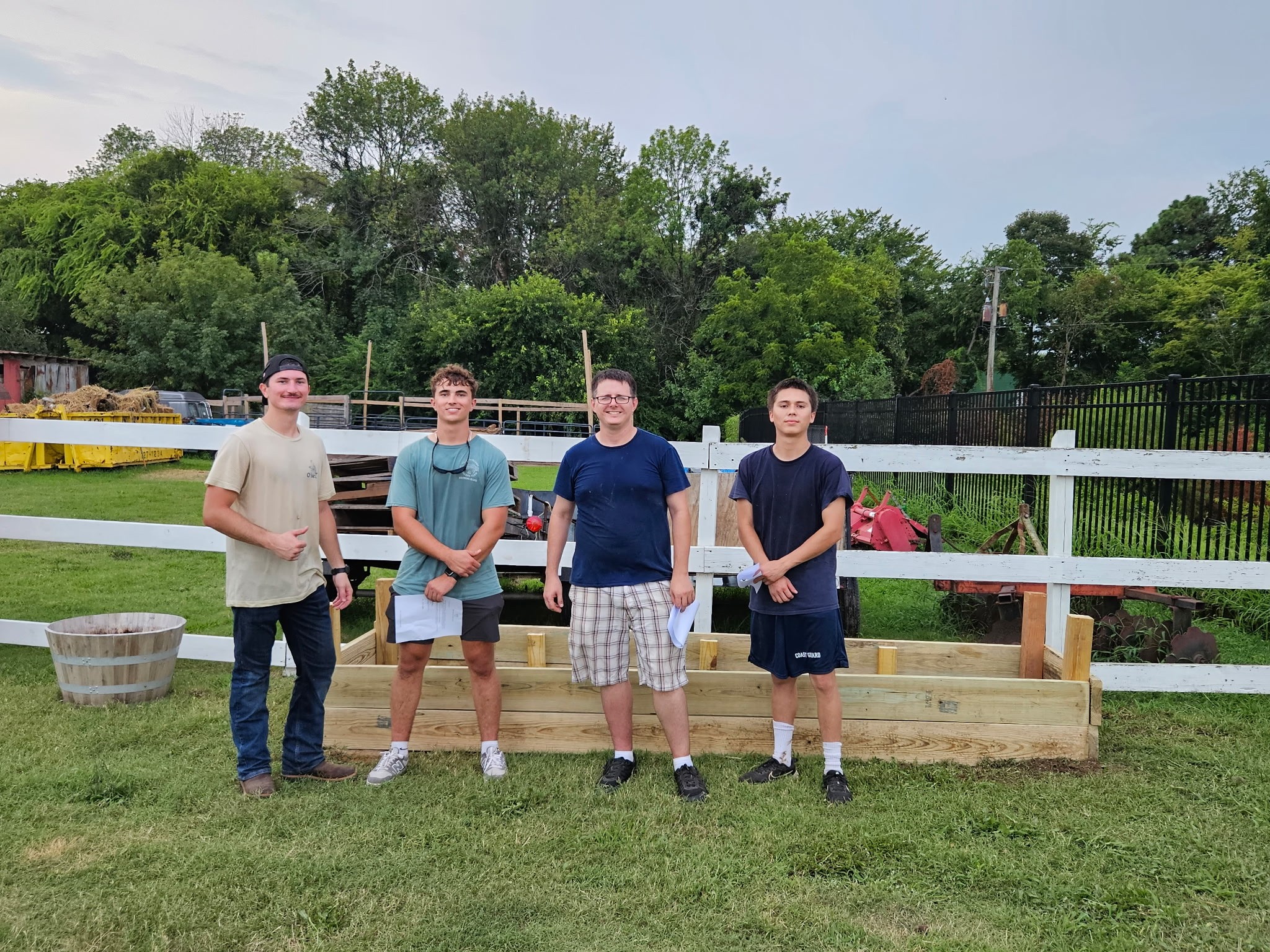 Four people stand in front of a wooden garden bed and white fence, with trees behind them.