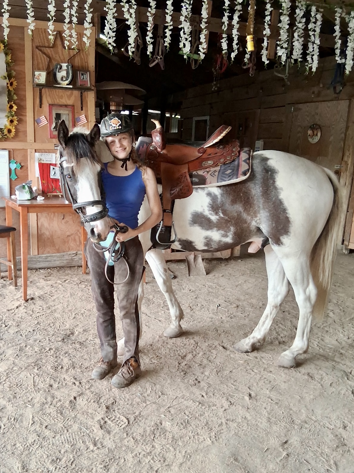 A person wearing a helmet stands beside a saddled brown-and-white horse in a stable.