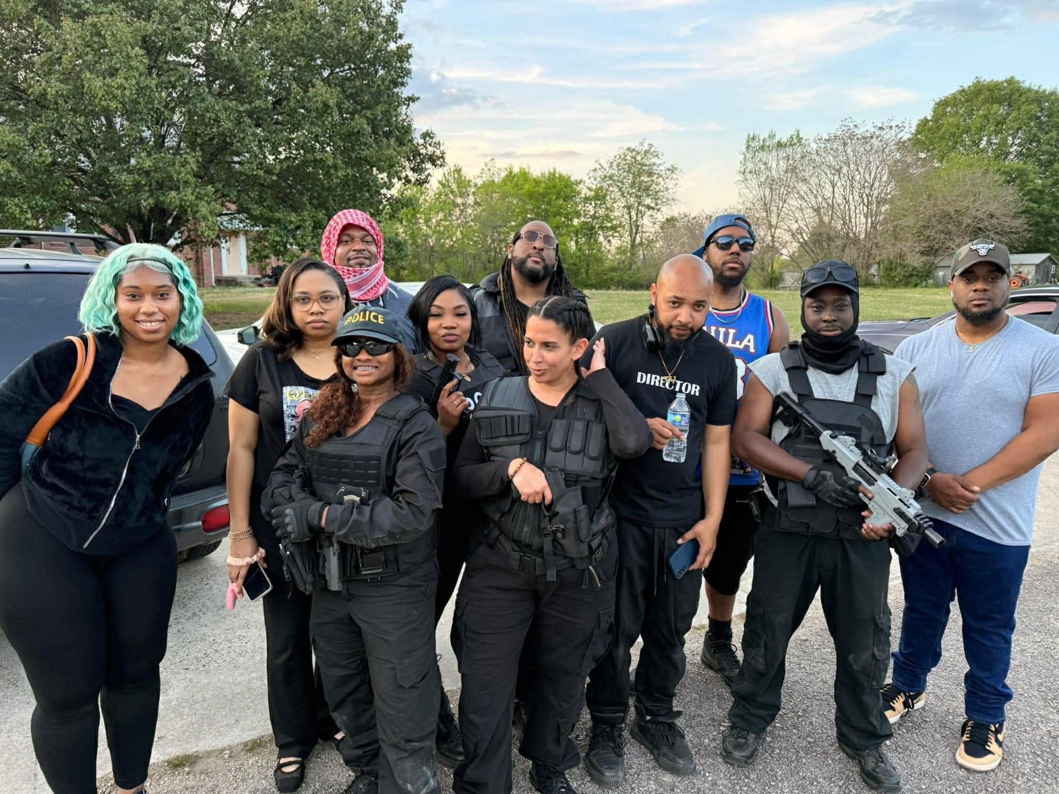 Group of ten people standing outdoors, some in tactical gear, with trees and a car in the background.