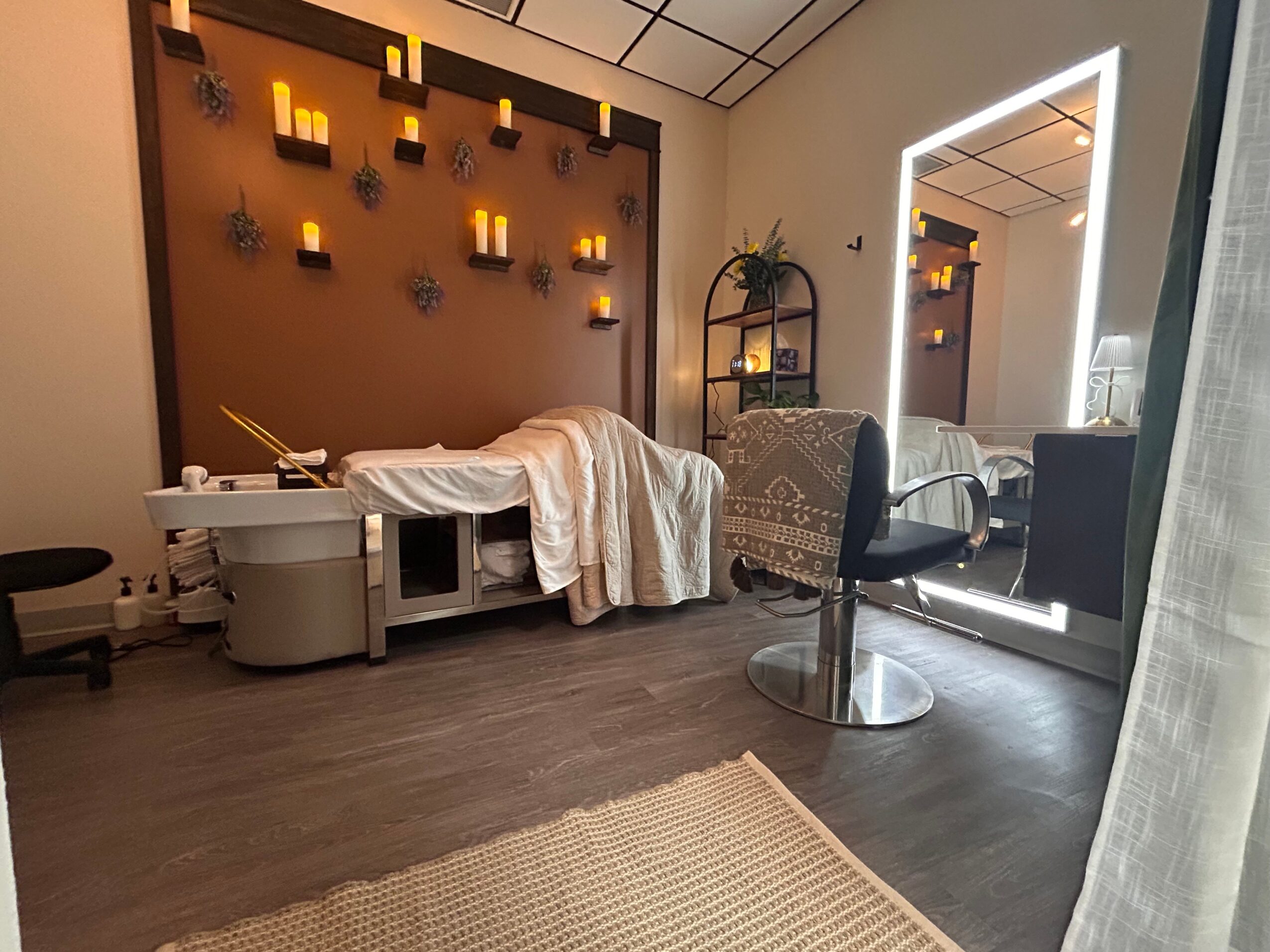 Massage table with a blanket, candles on wall, and a mirror in a spa room, with a chair and a large mirror.