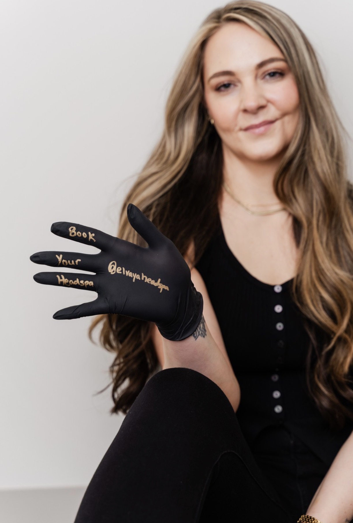 Young woman with long wavy hair holding a black glove with gold writing, smiling softly, wearing a black top, against a plain background.