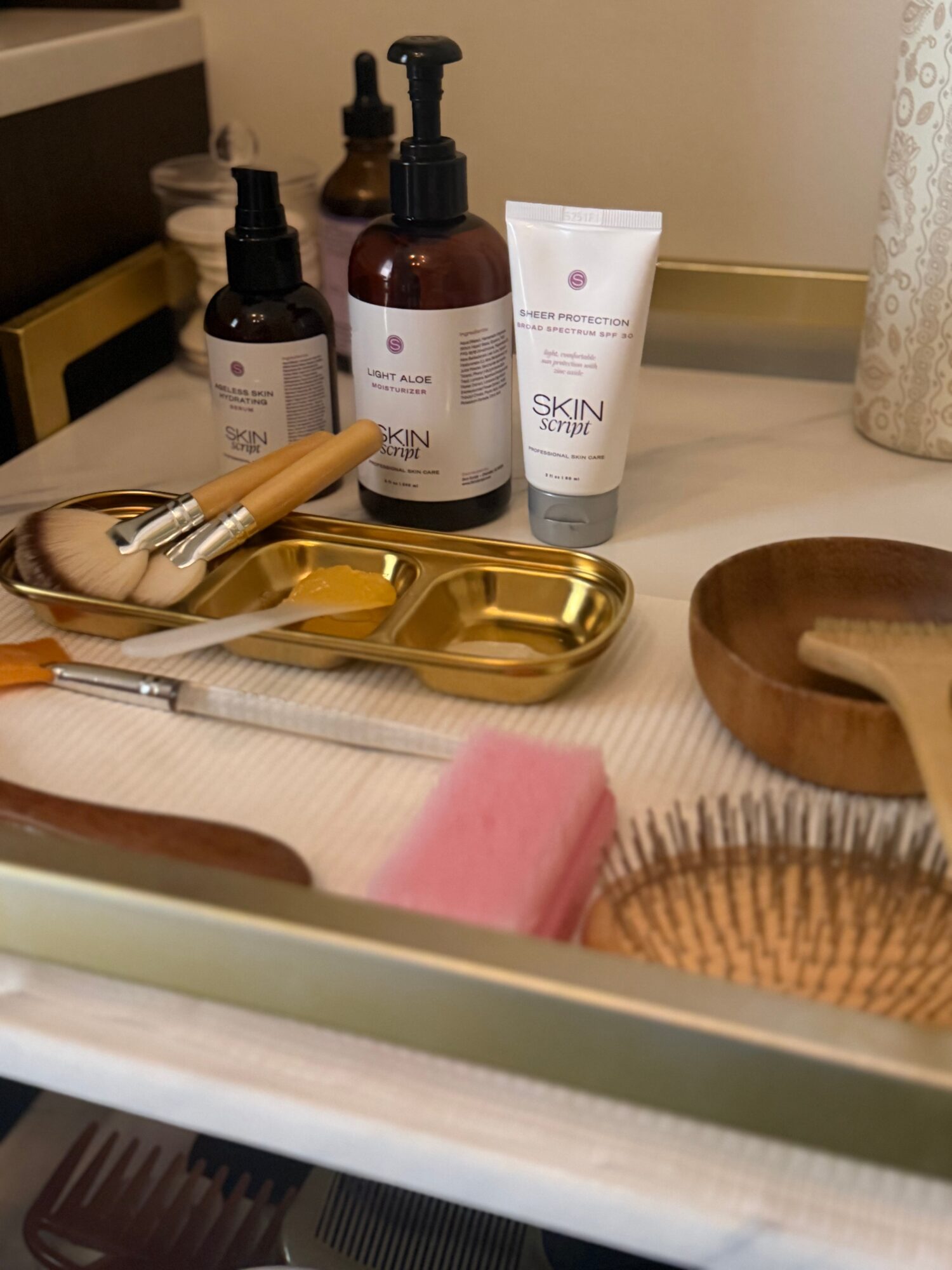 Skincare products, brushes, and a pink sponge on a bathroom countertop with a wooden bowl and a gold tray.