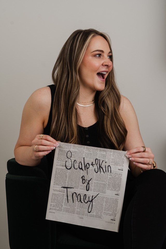 Woman with long hair holding a newspaper, smiling and looking to the side, against a plain background.