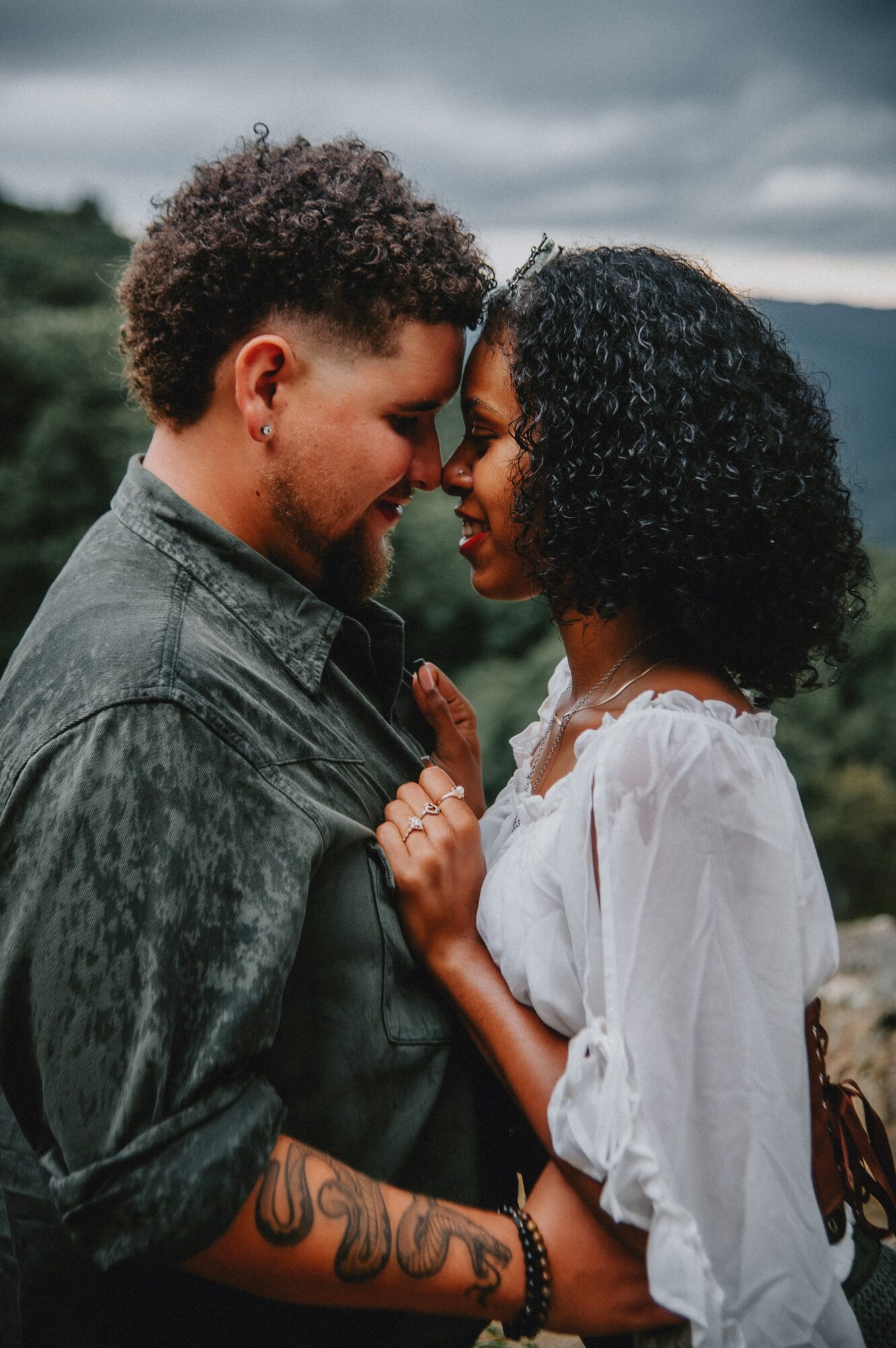 A couple faces close together outdoors, touching foreheads, with trees and cloudy sky in background.