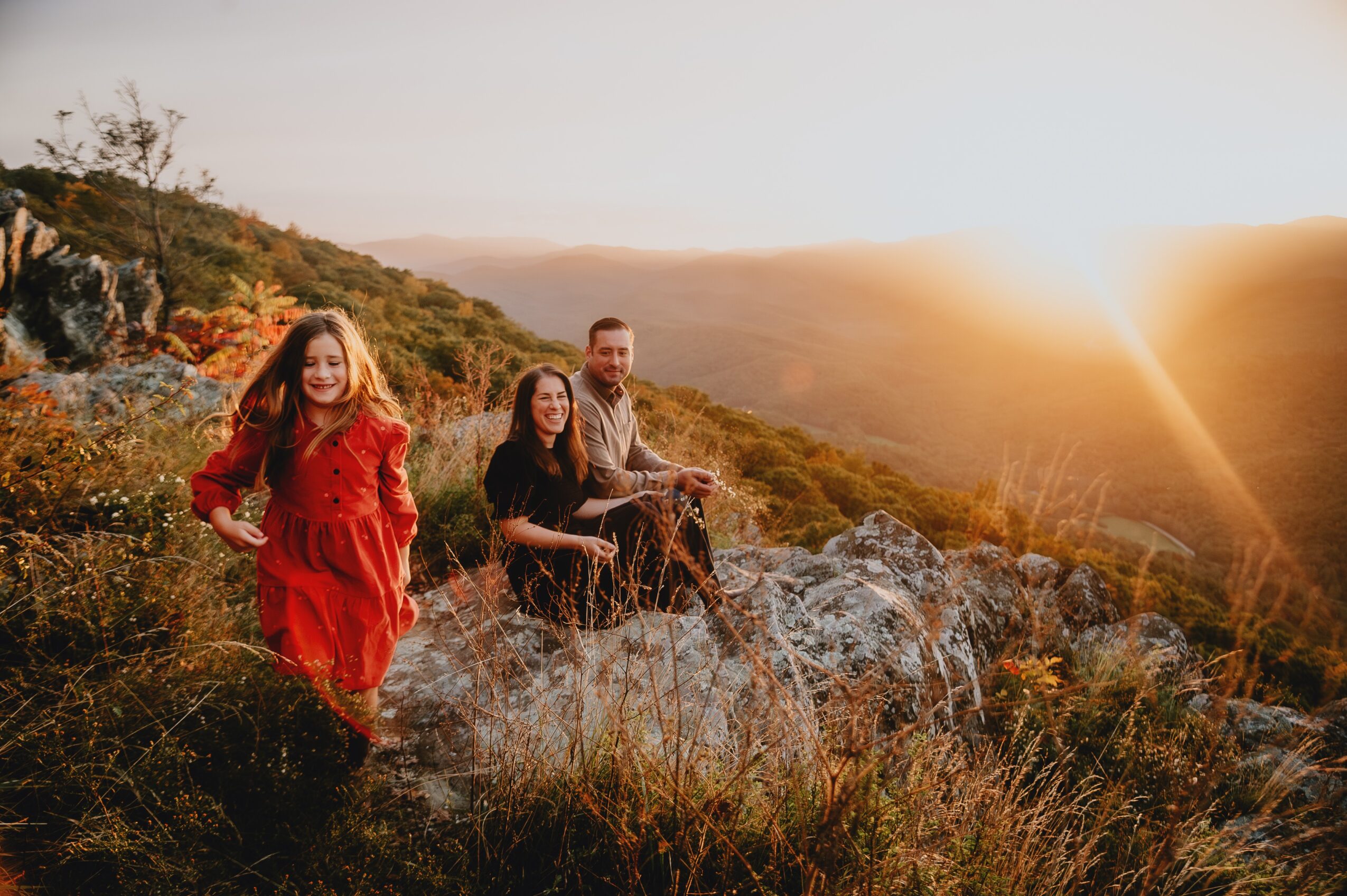 Three people sitting on rocks outdoors during sunset, surrounded by grass and hills, with a bright sky and warm light.