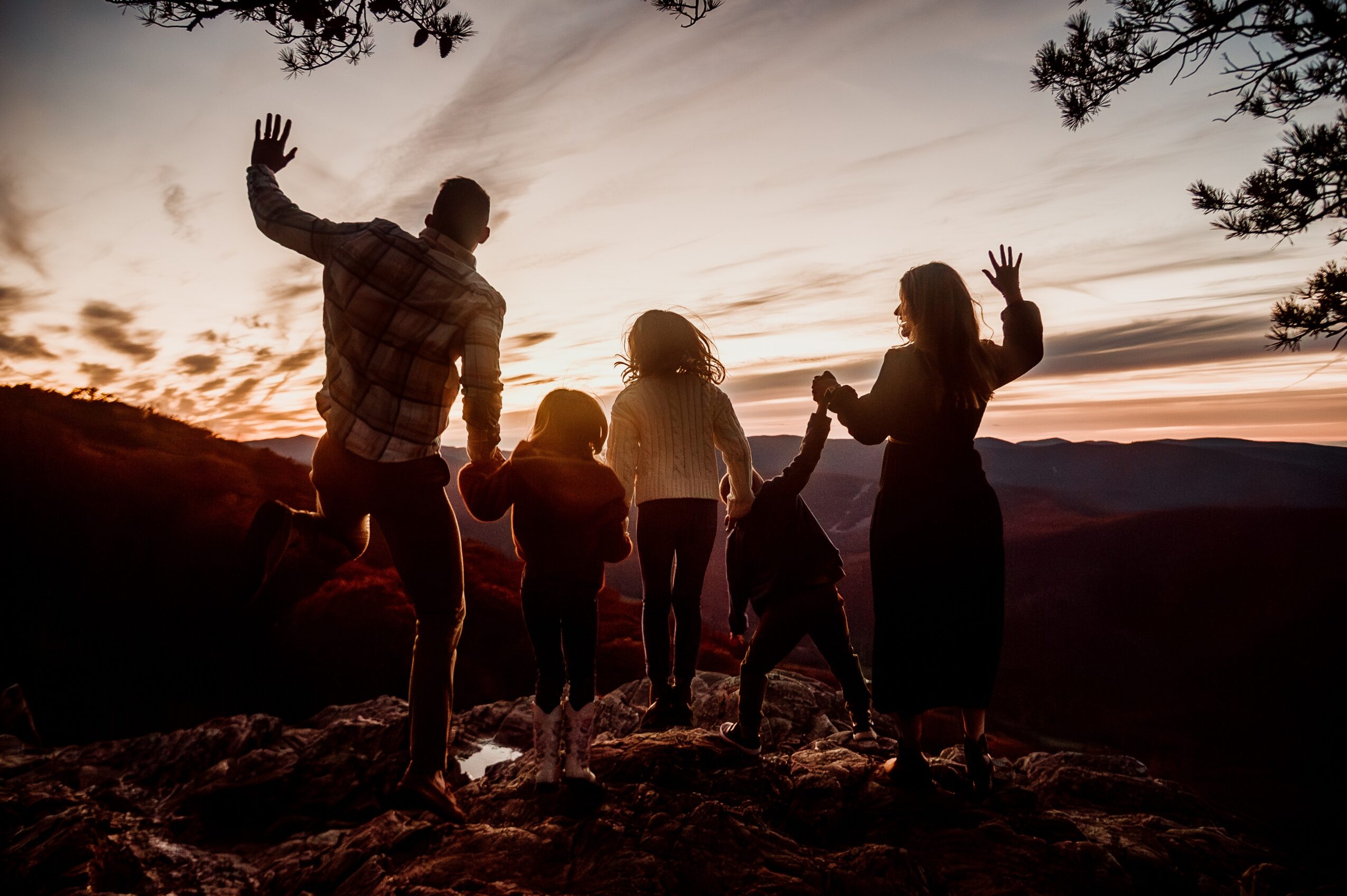 Silhouette of a family holding hands and waving at sunset on a rocky landscape with trees and a cloudy sky.