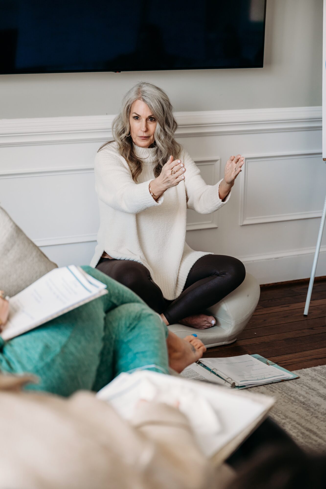 Woman with gray hair sitting on the floor, gesturing with hands during discussion, in a room with a large screen and books.