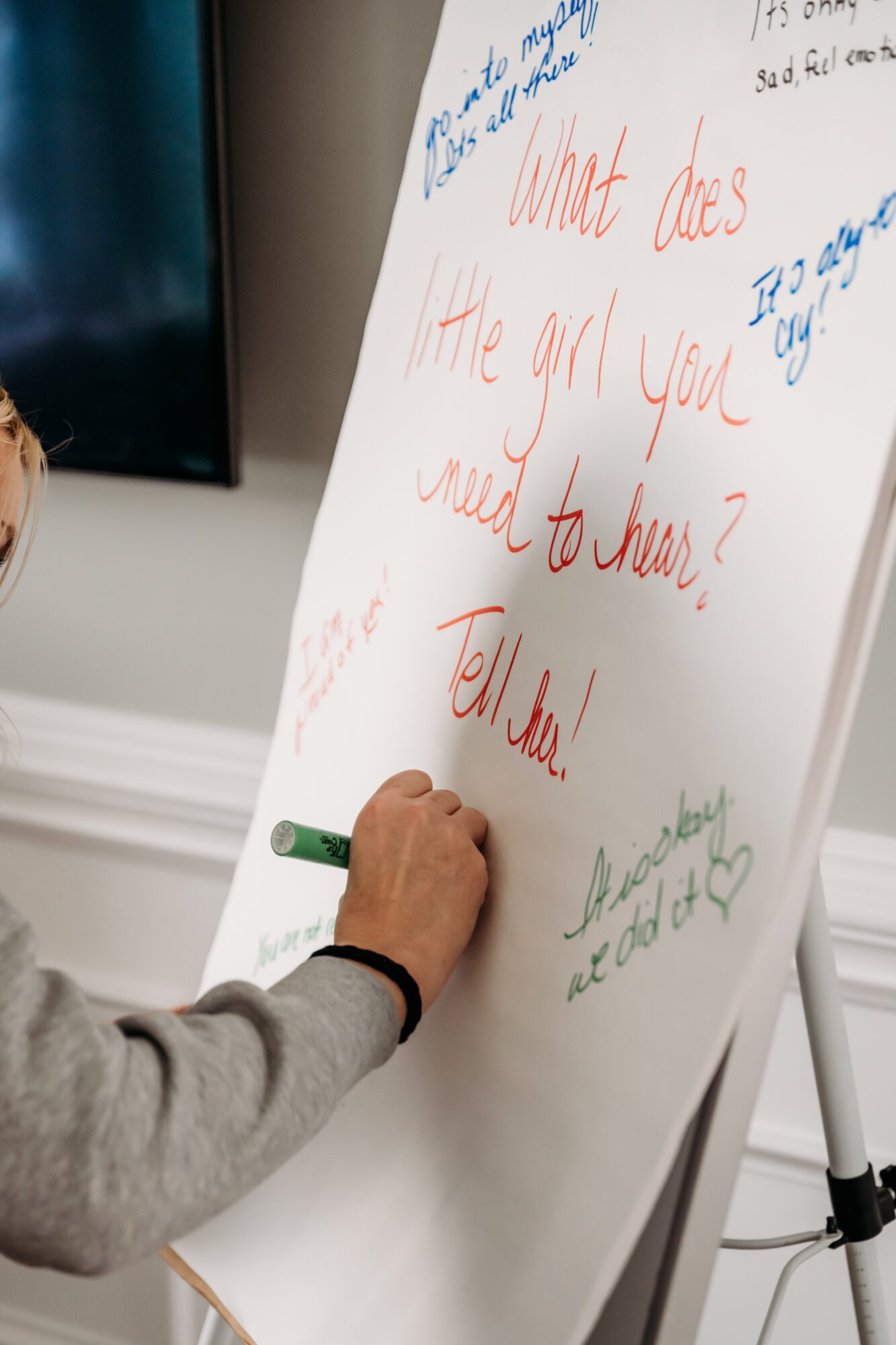 Person writing on a whiteboard with red and blue marker, whiteboard on a stand, hand visible, gray sleeve, room setting.